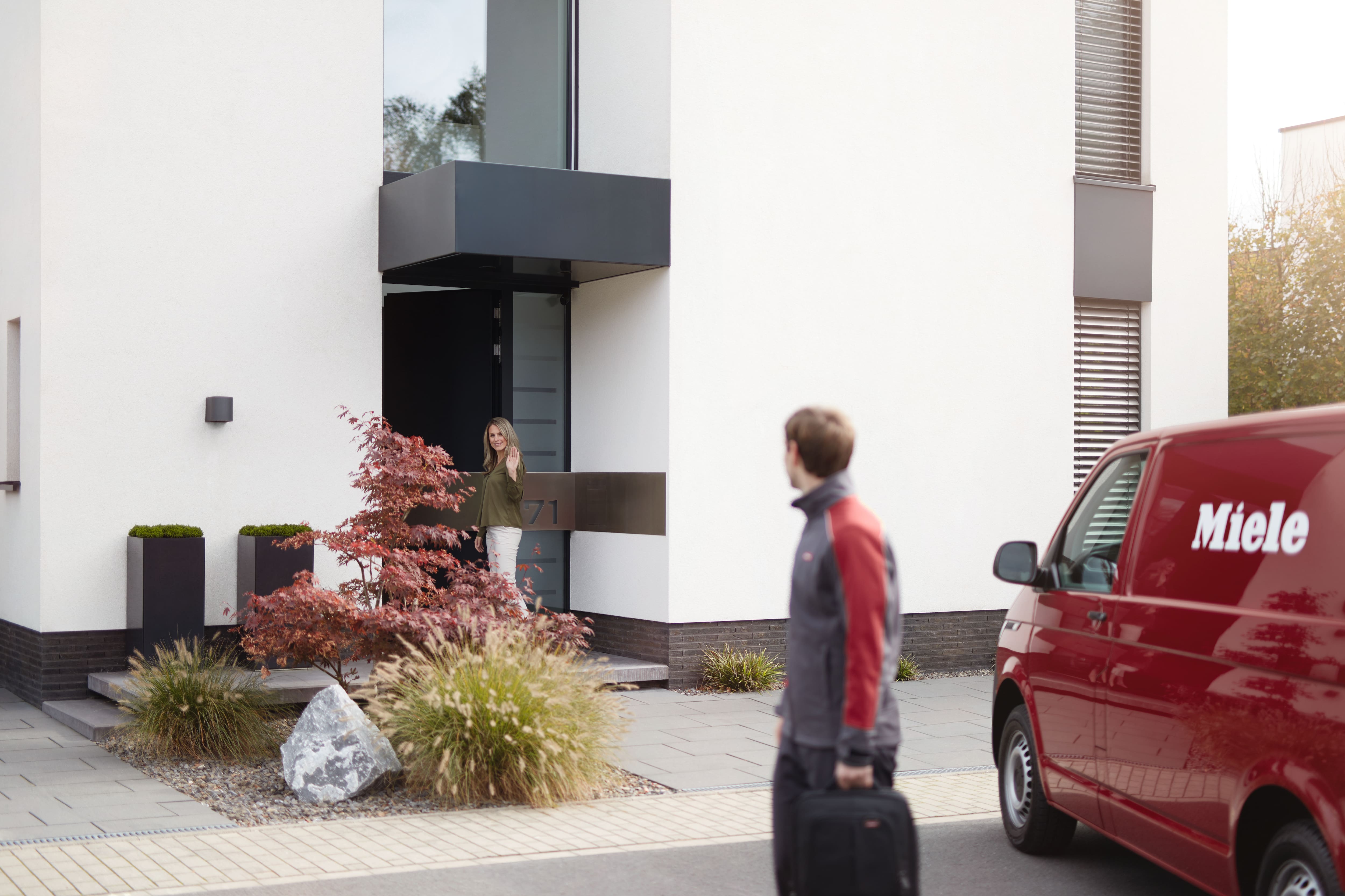 Technician approaches modern home as a person waves from the doorway; a red Miele service van is parked nearby.