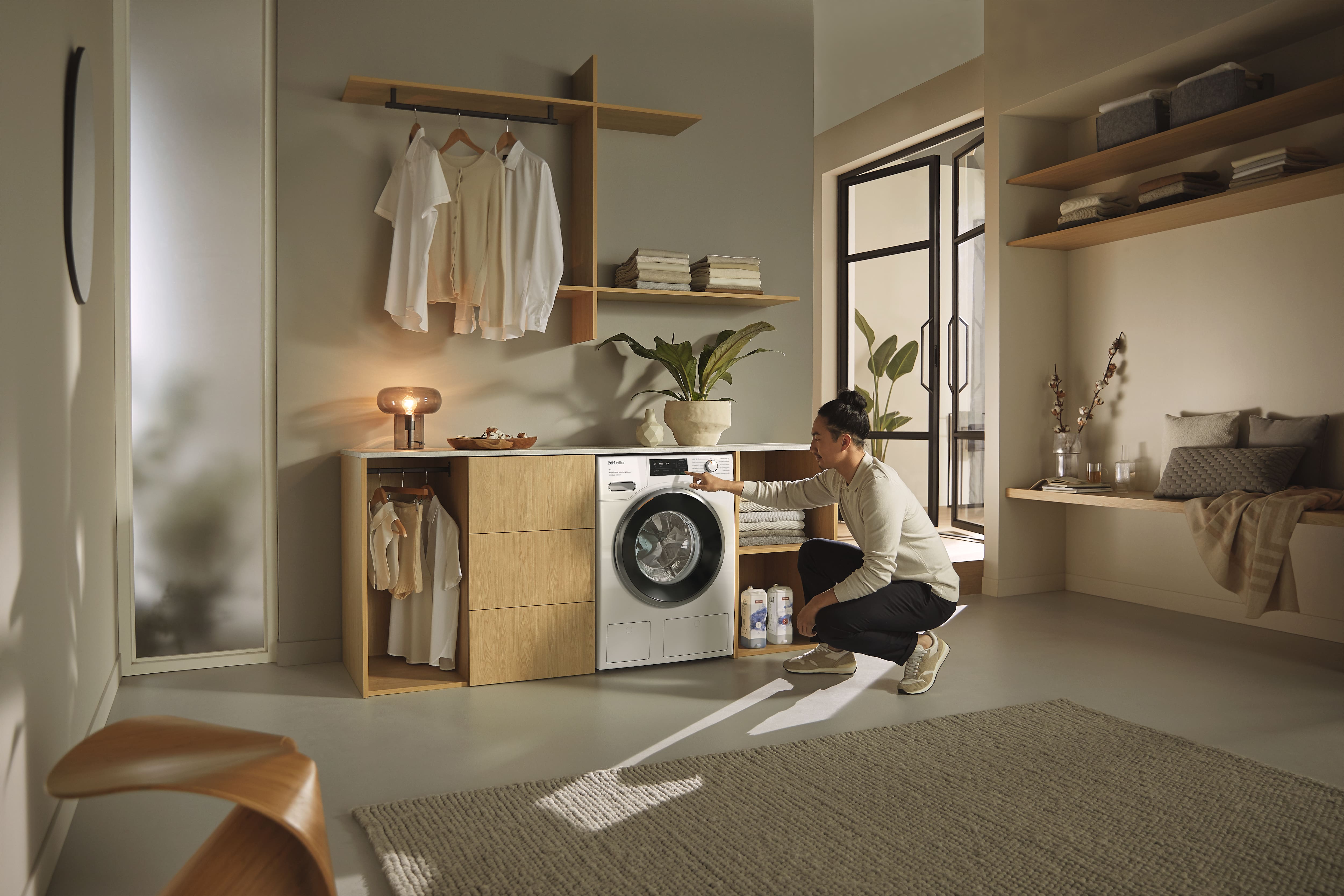 Person, der die Einstellungen an einer Frontlader-Waschmaschine in einem modernen, minimalistischen Waschraum mit Holzregalen, aufgehängter heller Kleidung, Zimmerpflanzen und nahegelegenen Waschmittelflaschen anpasst. Person, der die Einstellungen an einer Frontlader-Waschmaschine in einem modernen, minimalistischen Waschraum mit Holzregalen, aufgehängter heller Kleidung, Zimmerpflanzen und nahegelegenen Waschmittelflaschen anpasst.