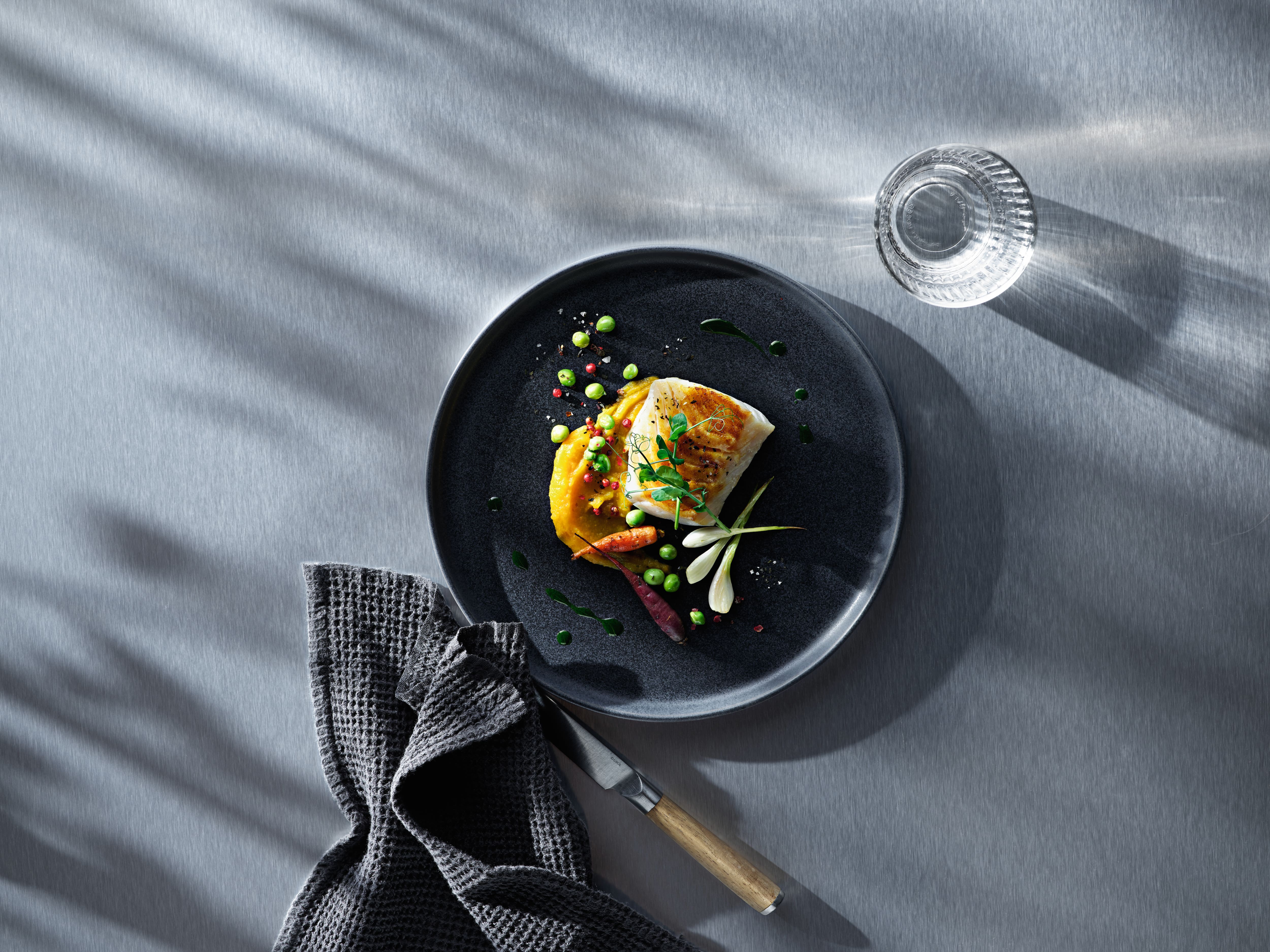 Top-down view of seared fish with puree and peas on a dark plate, with towel, knife, and glass of water on a gray table. Top-down view of seared fish with puree and peas on a dark plate, with towel, knife, and glass of water on a gray table.