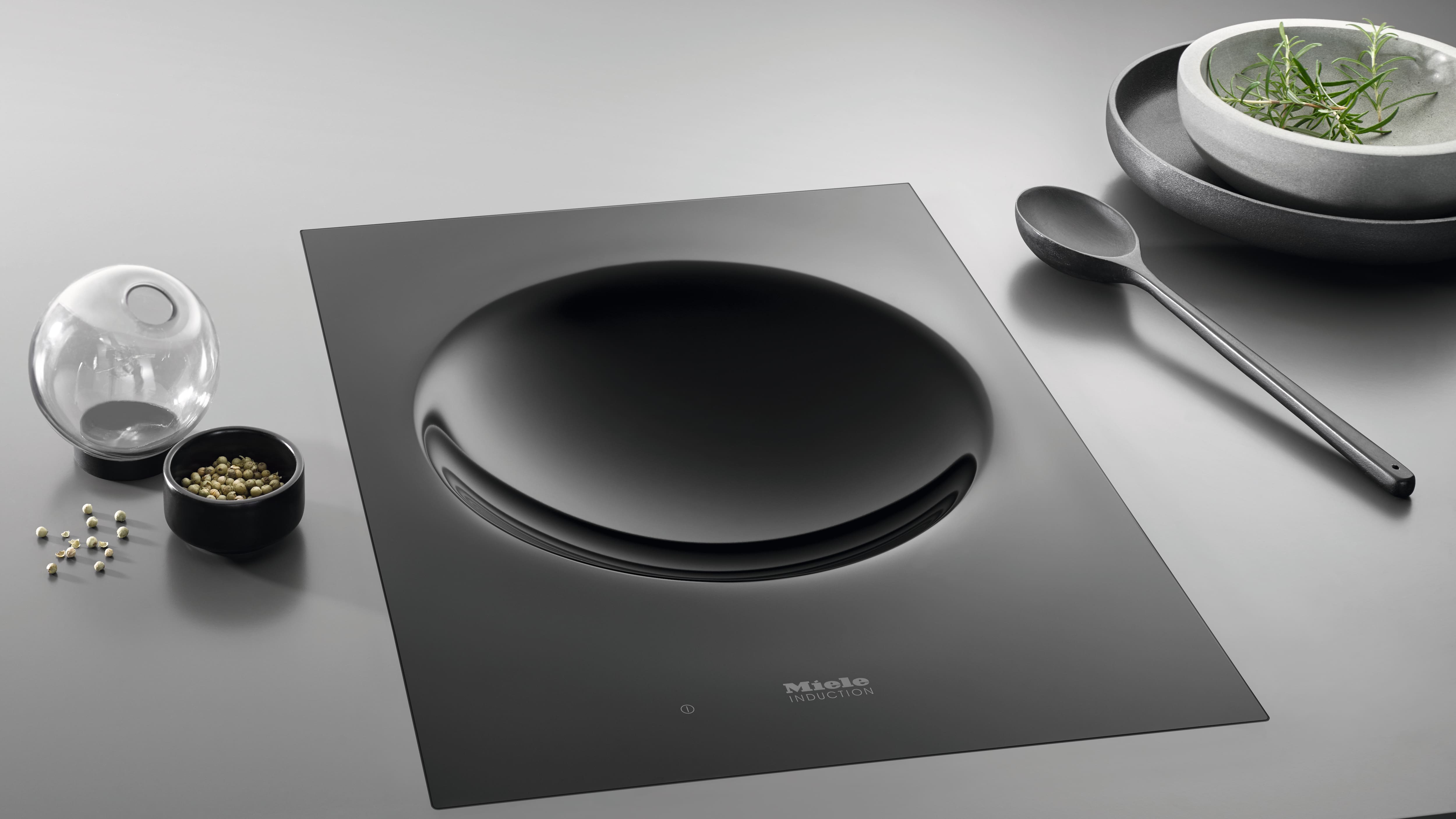 Sleek black Miele induction cooktop with a concave wok zone, shown on a minimalist countertop alongside a spoon, bowls, herbs, and peppercorns. Sleek black Miele induction cooktop with a concave wok zone, shown on a minimalist countertop alongside a spoon, bowls, herbs, and peppercorns.