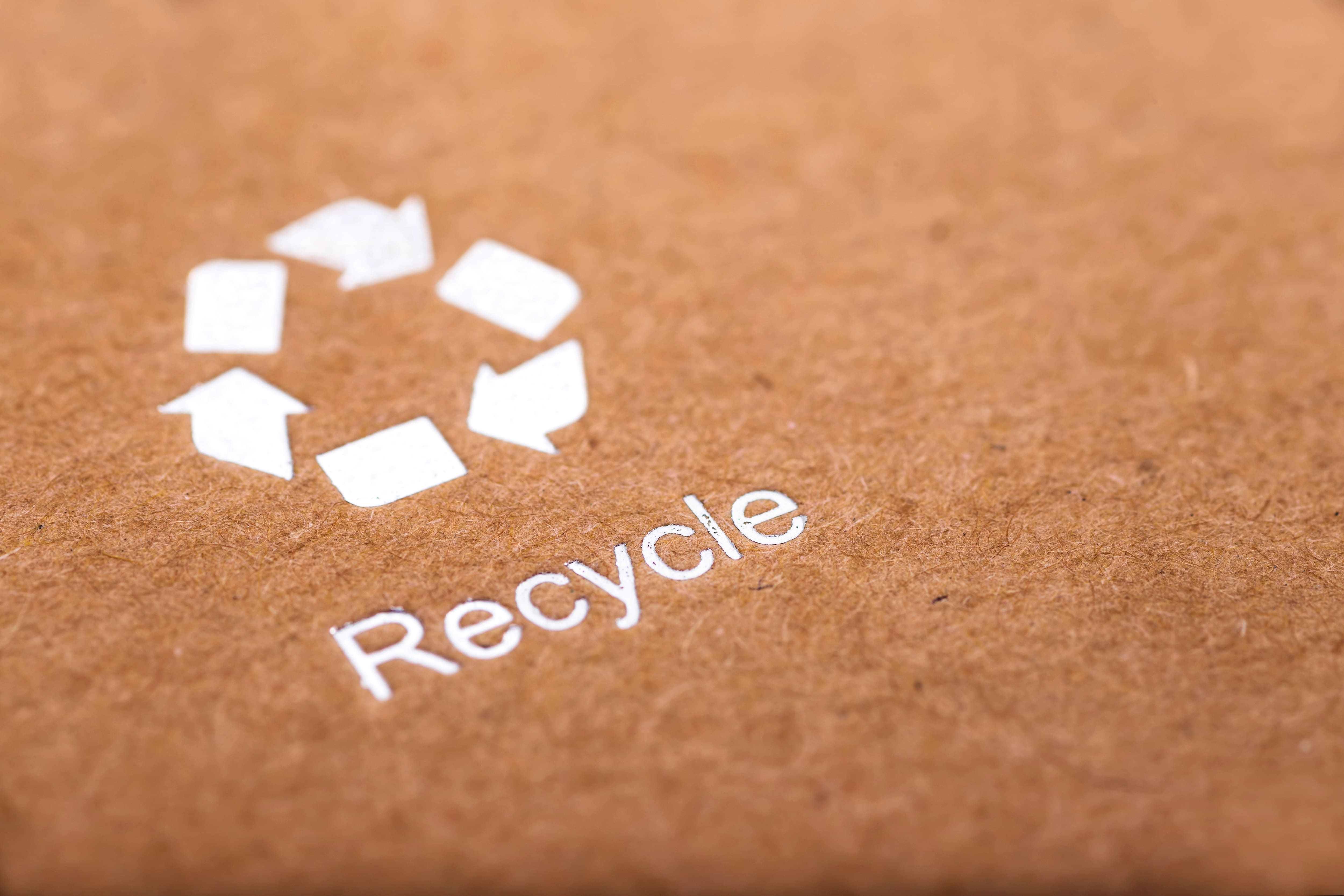 White recycle symbol and the word “Recycle” printed on brown kraft paper, highlighting eco-friendly packaging. White recycle symbol and the word “Recycle” printed on brown kraft paper, highlighting eco-friendly packaging.
