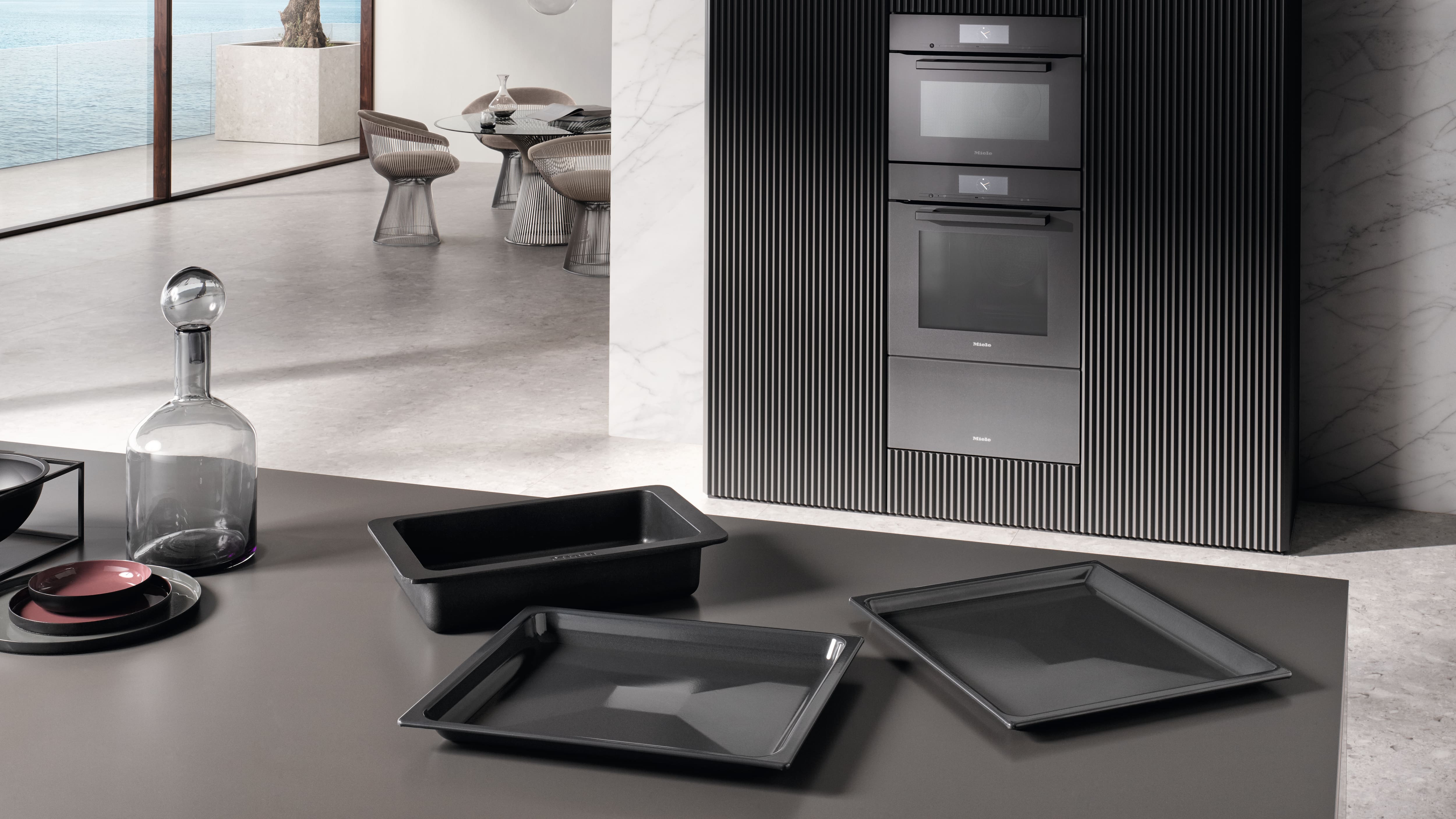 Cucina moderna e minimalista con vista sul mare, forni neri integrati in un frontale scanalato e dettagli in marmo; in primo piano, un piano di lavoro scuro con teglie da forno, una caraffa in vetro e piatti impilati. Cucina moderna e minimalista con vista sul mare, forni neri integrati in un frontale scanalato e dettagli in marmo; in primo piano, un piano di lavoro scuro con teglie da forno, una caraffa in vetro e piatti impilati.
