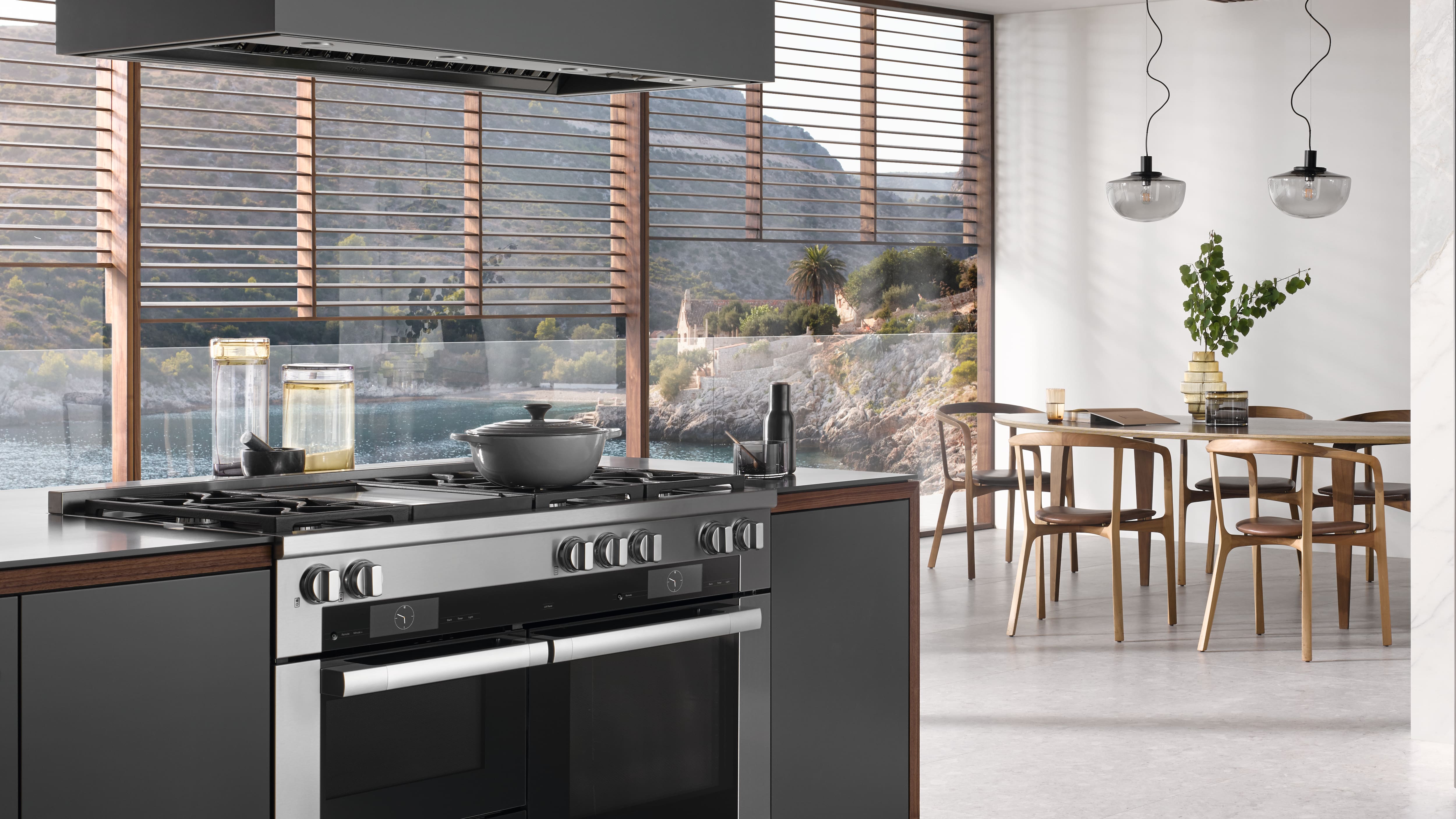 Modern open kitchen and dining area with a stainless steel gas range and lidded pot in the foreground, sleek dark cabinetry, pendant lights, and floor‑to‑ceiling windows with wooden slats overlooking a calm coastal bay with rocky Modern open kitchen and dining area with a stainless steel gas range and lidded pot in the foreground, sleek dark cabinetry, pendant lights, and floor‑to‑ceiling windows with wooden slats overlooking a calm coastal bay with rocky