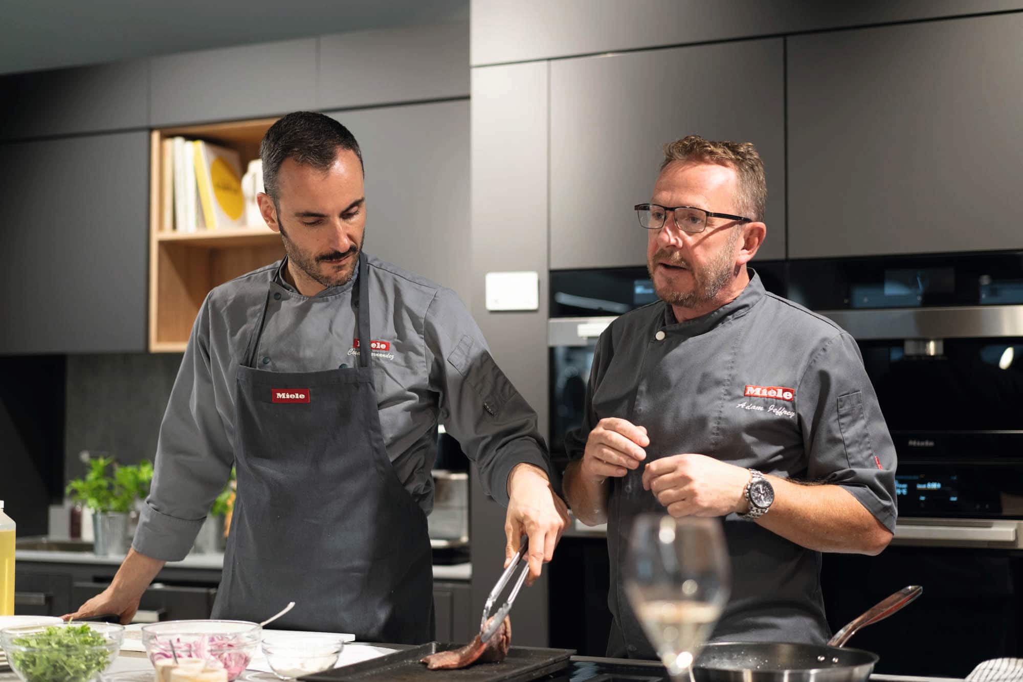 Two chefs in grey Miele uniforms lead a cooking demo, searing meat with tongs in a modern kitchen studio.