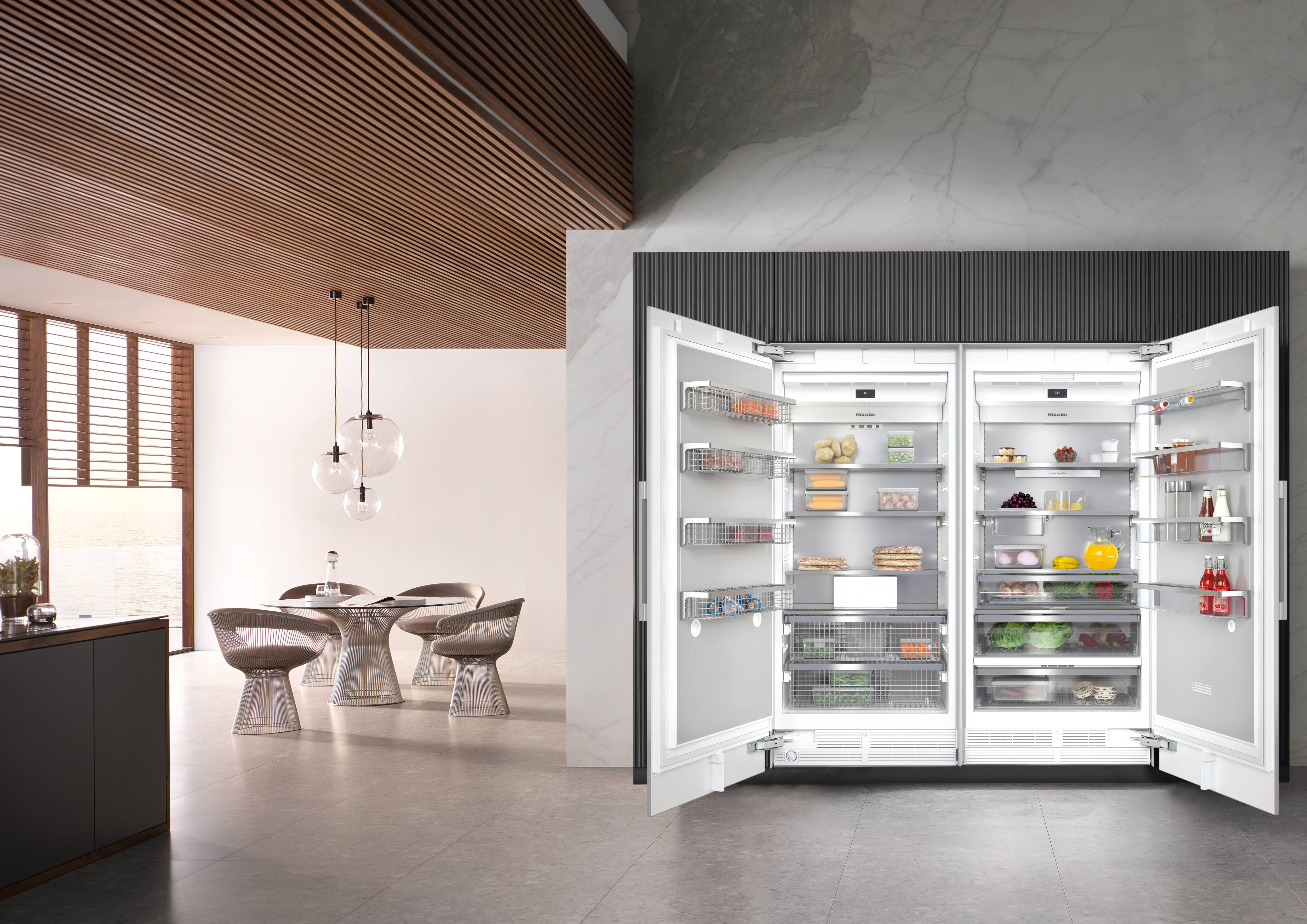 Cozinha moderna com teto de ripas de madeira e parede de mármore, com uma geladeira embutida dupla aberta, repleta de alimentos, ao lado de uma mesa de jantar redonda com quatro cadeiras de malha e luminárias pendentes em forma de globo. Cozinha moderna com teto de ripas de madeira e parede de mármore, com uma geladeira embutida dupla aberta, repleta de alimentos, ao lado de uma mesa de jantar redonda com quatro cadeiras de malha e luminárias pendentes em forma de globo.