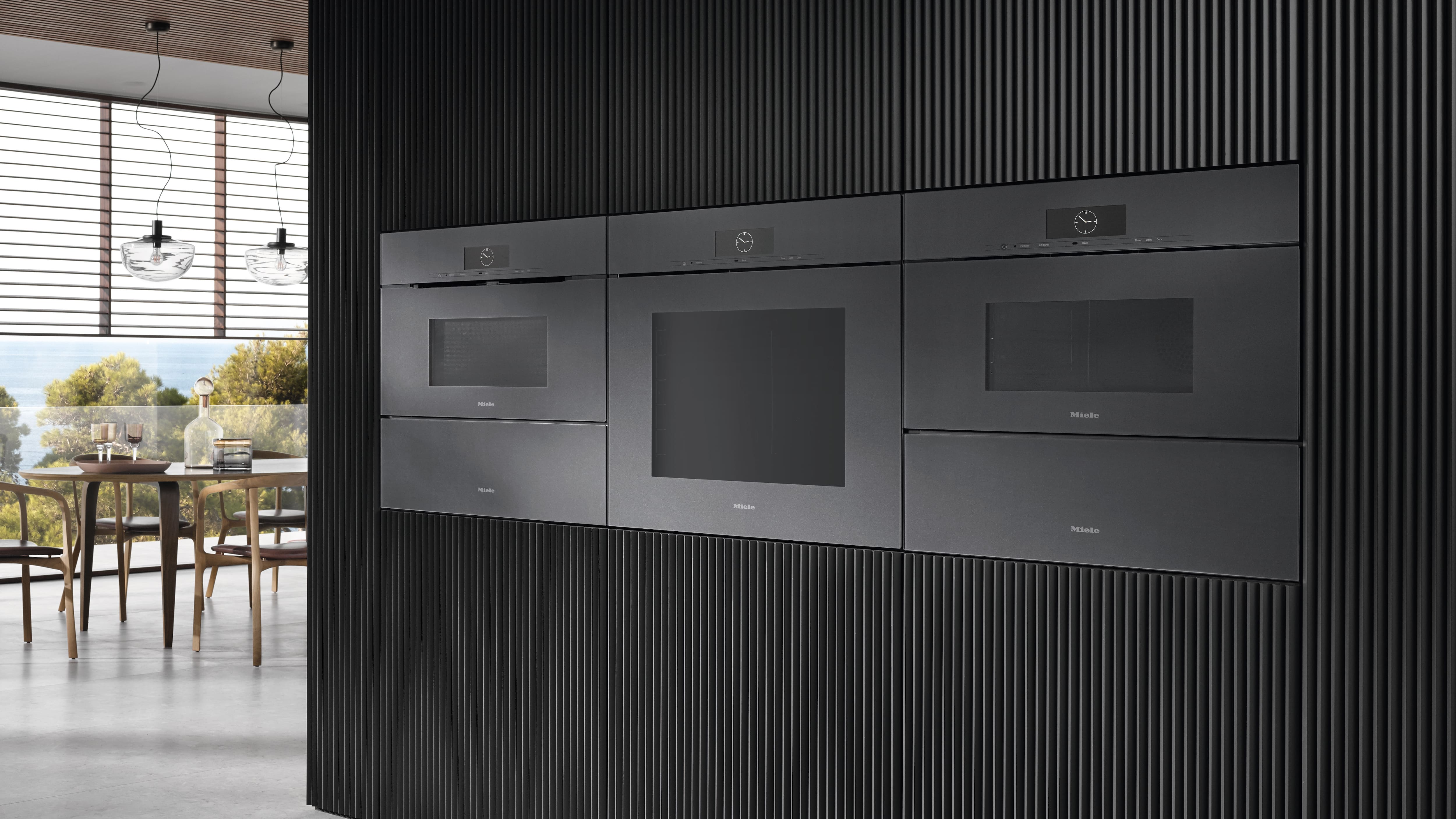 Sleek black built-in Miele ovens and warming drawers in a ribbed wall, with airy dining area and scenic view beyond.
