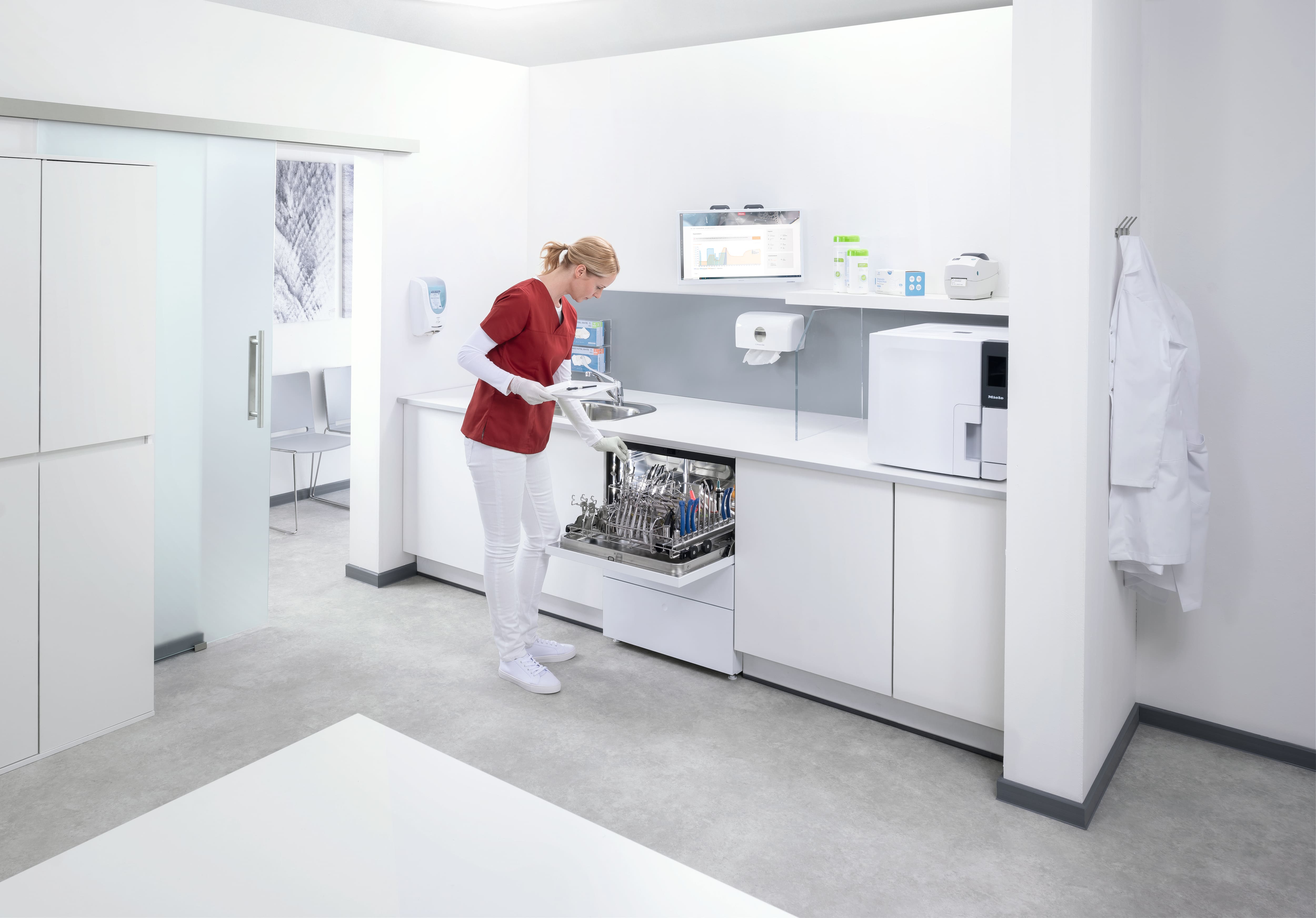 Sjukvårdspersonal i röda operationskläder diskar instrument av metall för medicinskt och dentalt bruk i en steriliseringsmaskin i ett rent, vitt rum med skåp och utrustning för rengöring på en klinik.