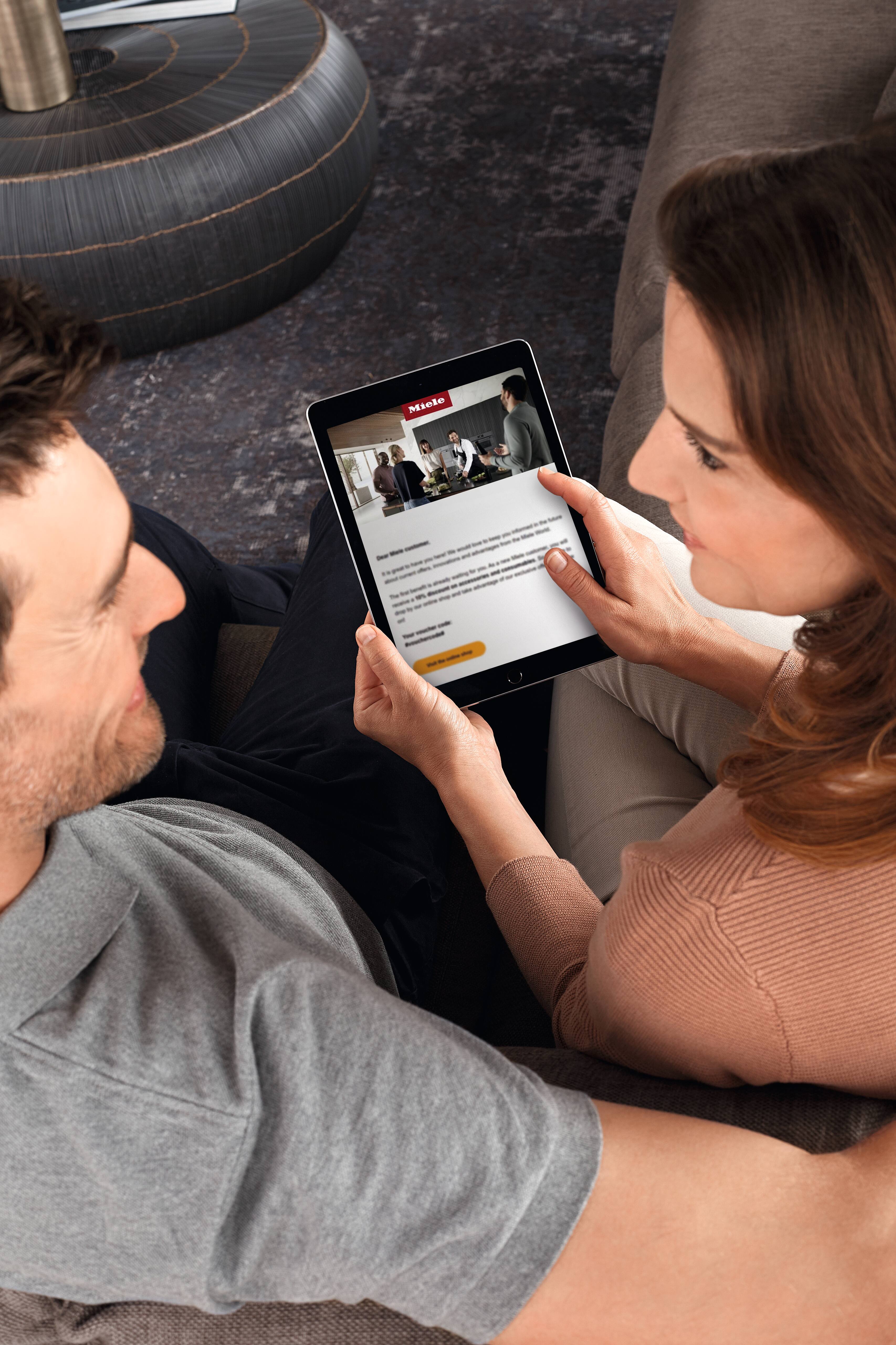 Um casal sentado no sofá observa um tablet que exibe uma página da Miele com uma cena de culinária e um botão de inscrição. Um casal sentado no sofá observa um tablet que exibe uma página da Miele com uma cena de culinária e um botão de inscrição.