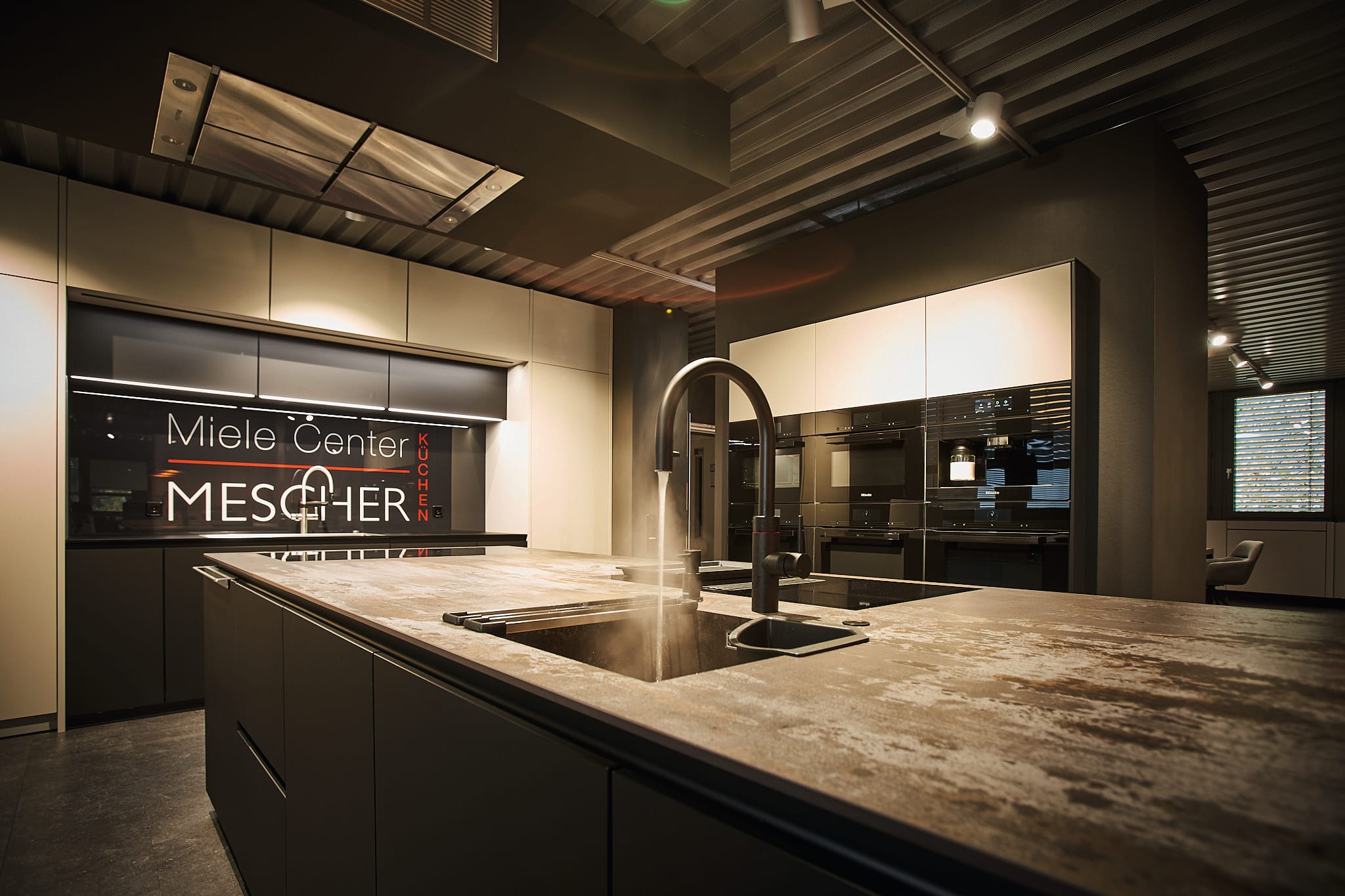 Moderne Showroom-Küche mit einer großen Insel, die eine Arbeitsplatte im Betonstil, ein Spülbecken mit heißem Wasser aus einem hohen Wasserhahn, matte dunkle Schränke, eingebaute schwarze Miele-Geräte an der Rückwand, industrielle Deckenbeleuchtung und ein Schild mit der Aufschrift „Miele Center Mescher Küchen“ im Hintergrund bietet.