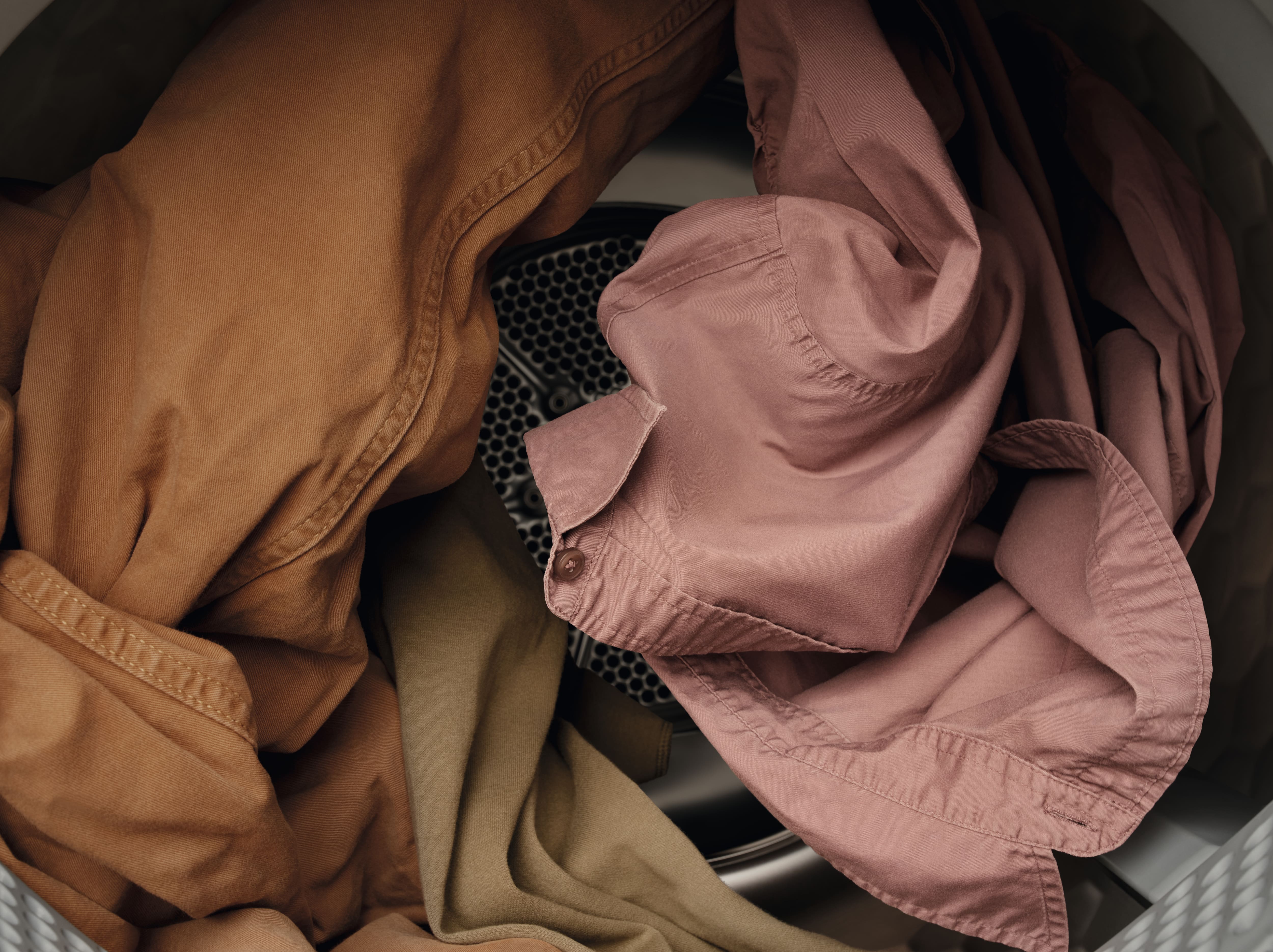 Camisas en tonos tierra y tela en el interior del tambor de una lavadora, en las que se aprecian prendas de color rosa, marrón y verde oliva en pleno proceso de lavado. Camisas en tonos tierra y tela en el interior del tambor de una lavadora, en las que se aprecian prendas de color rosa, marrón y verde oliva en pleno proceso de lavado.