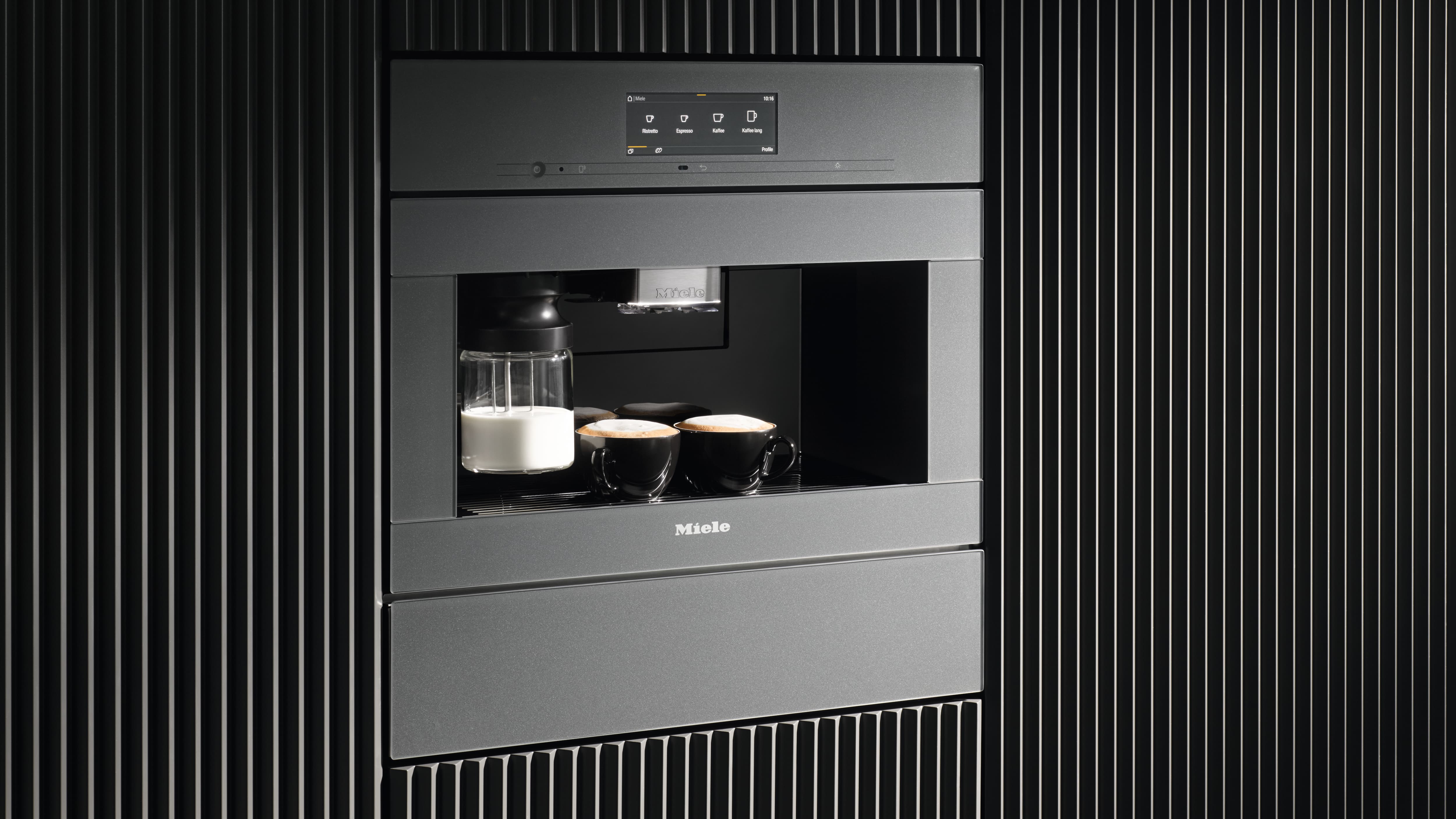 Built-in Miele coffee machine with touchscreen, milk container, and two cappuccinos in a sleek black paneled wall.