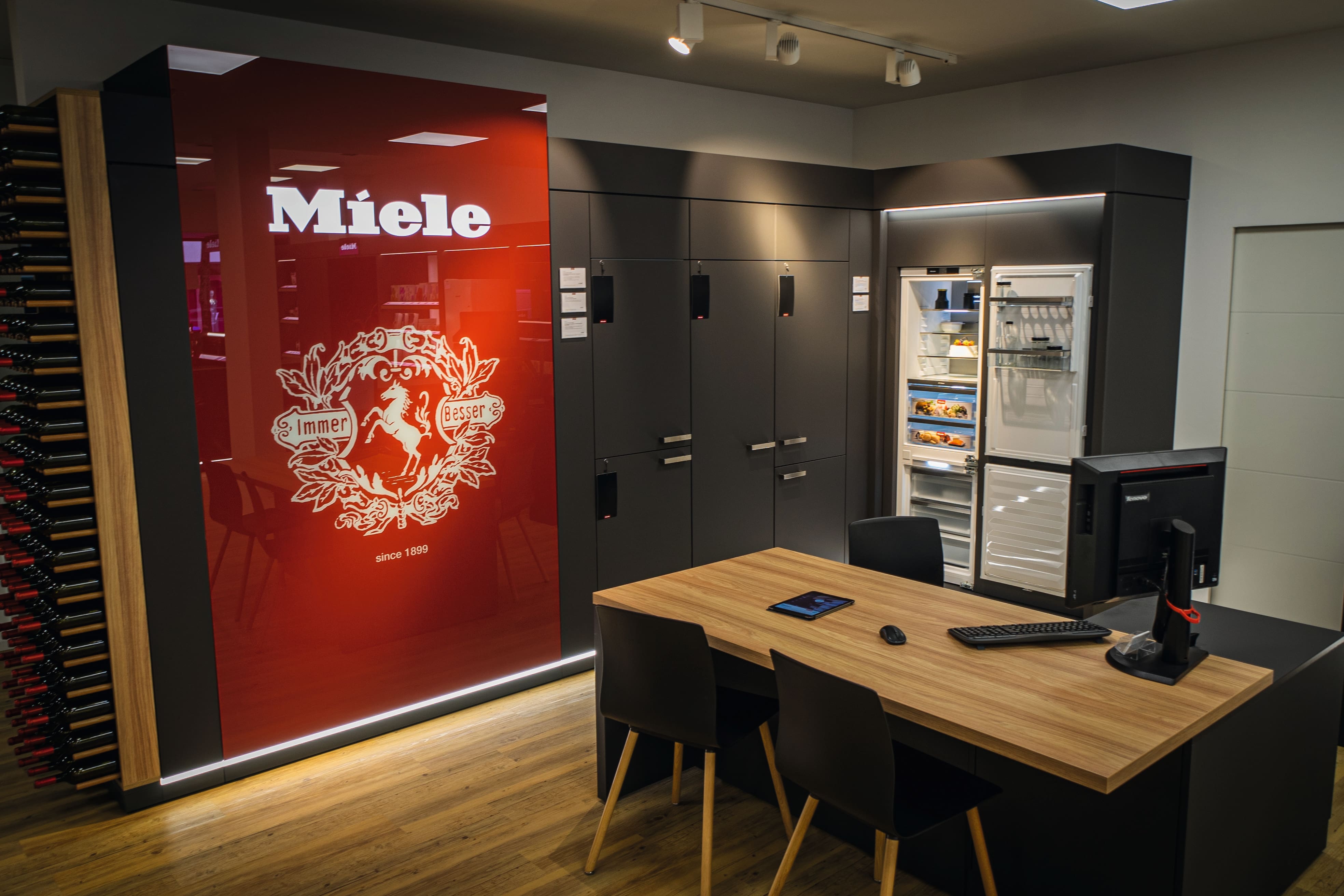 Moderner Miele-Geräteshowroom mit großer roter Markenwand, dunklen Einbauschränken, offenem Kühlschrank mit Lebensmitteln, einem Holzkonsultationstisch mit Computer und Tastatur sowie einem Weinregal links.