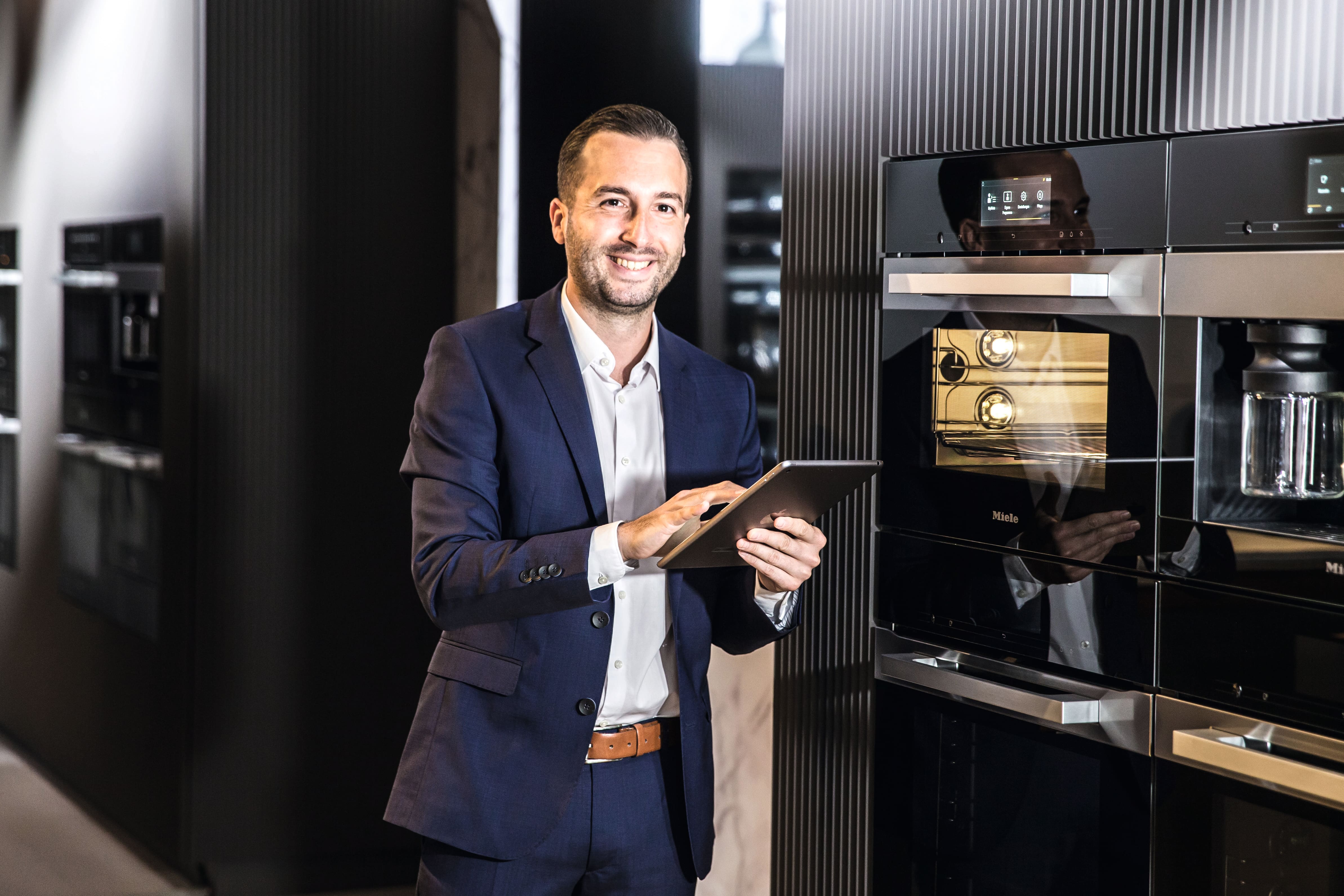 Geschäftsmann in einem marineblauen Anzug, der ein Tablet in einem modernen Küchengeräte-Showroom benutzt, neben eleganten Einbauöfen und einer Kaffeemaschine stehend. Geschäftsmann in einem marineblauen Anzug, der ein Tablet in einem modernen Küchengeräte-Showroom benutzt, neben eleganten Einbauöfen und einer Kaffeemaschine stehend.
