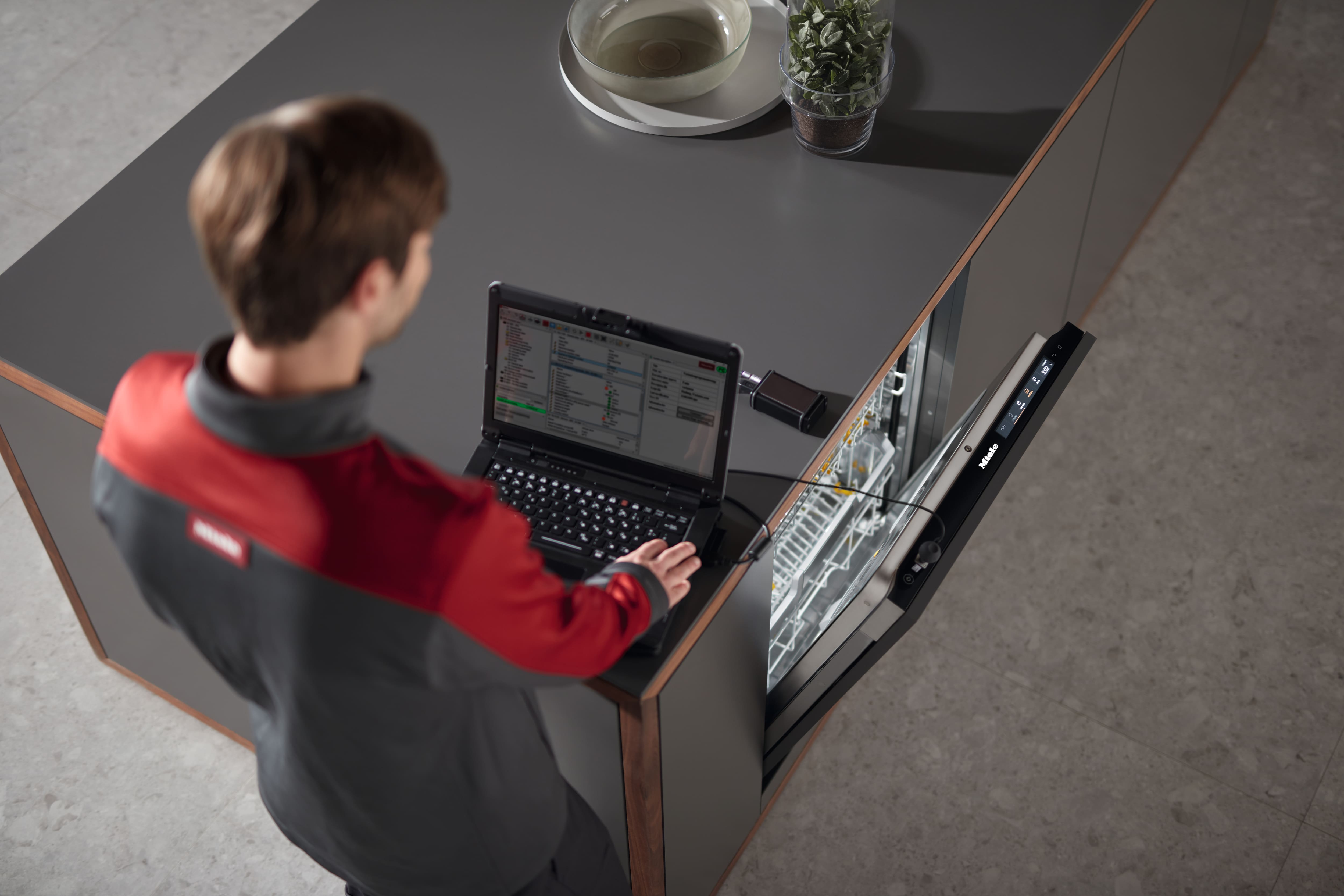 Ein Techniker führt mithilfe eines Laptops eine Diagnose an einem offenen Einbau-Geschirrspüler auf einer modernen Kücheninsel durch. Ein Techniker führt mithilfe eines Laptops eine Diagnose an einem offenen Einbau-Geschirrspüler auf einer modernen Kücheninsel durch.