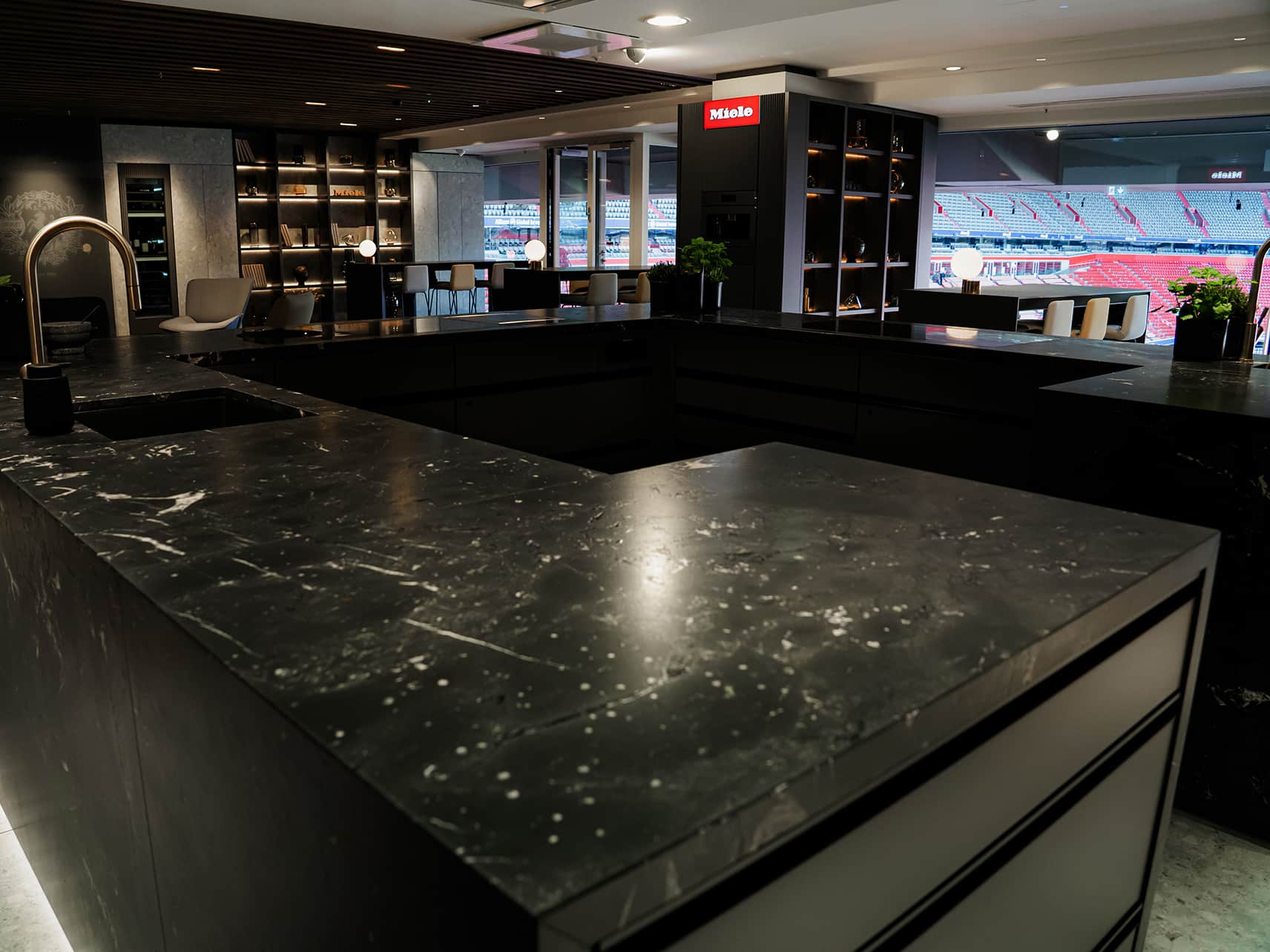 Moderne Luxus-Küchenlounge mit dunklen Marmorarbeitsflächen, Messingarmatur und offenen Regalen. Durch die großen Fenster bietet sich ein Blick auf das Stadion und verbindet elegantes Küchendesign mit exklusivem Ambiente. Moderne Luxus-Küchenlounge mit dunklen Marmorarbeitsflächen, Messingarmatur und offenen Regalen. Durch die großen Fenster bietet sich ein Blick auf das Stadion und verbindet elegantes Küchendesign mit exklusivem Ambiente.