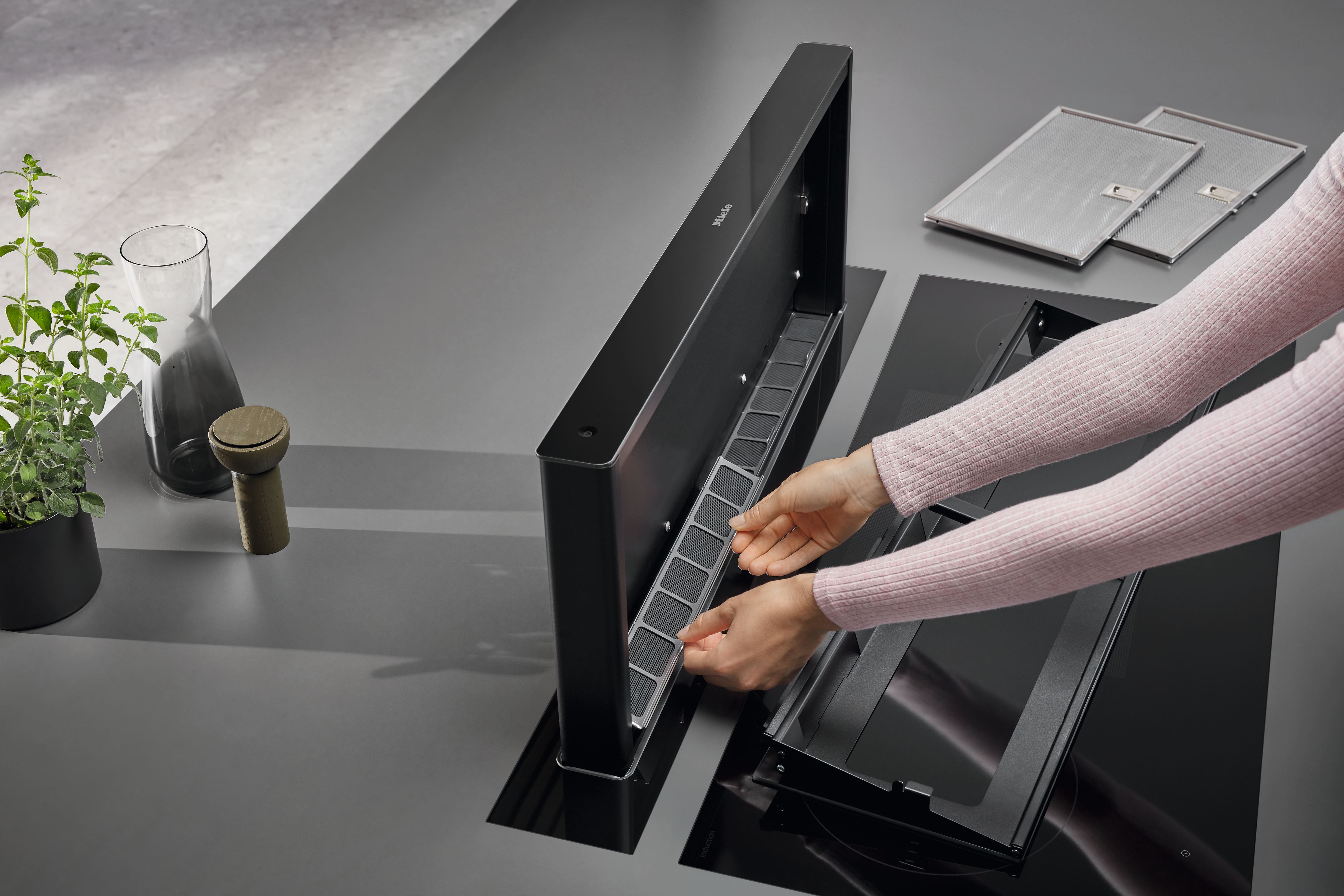 Hands replace a filter on a sleek pop-up downdraft kitchen extractor beside a glass cooktop and filters. Hands replace a filter on a sleek pop-up downdraft kitchen extractor beside a glass cooktop and filters.
