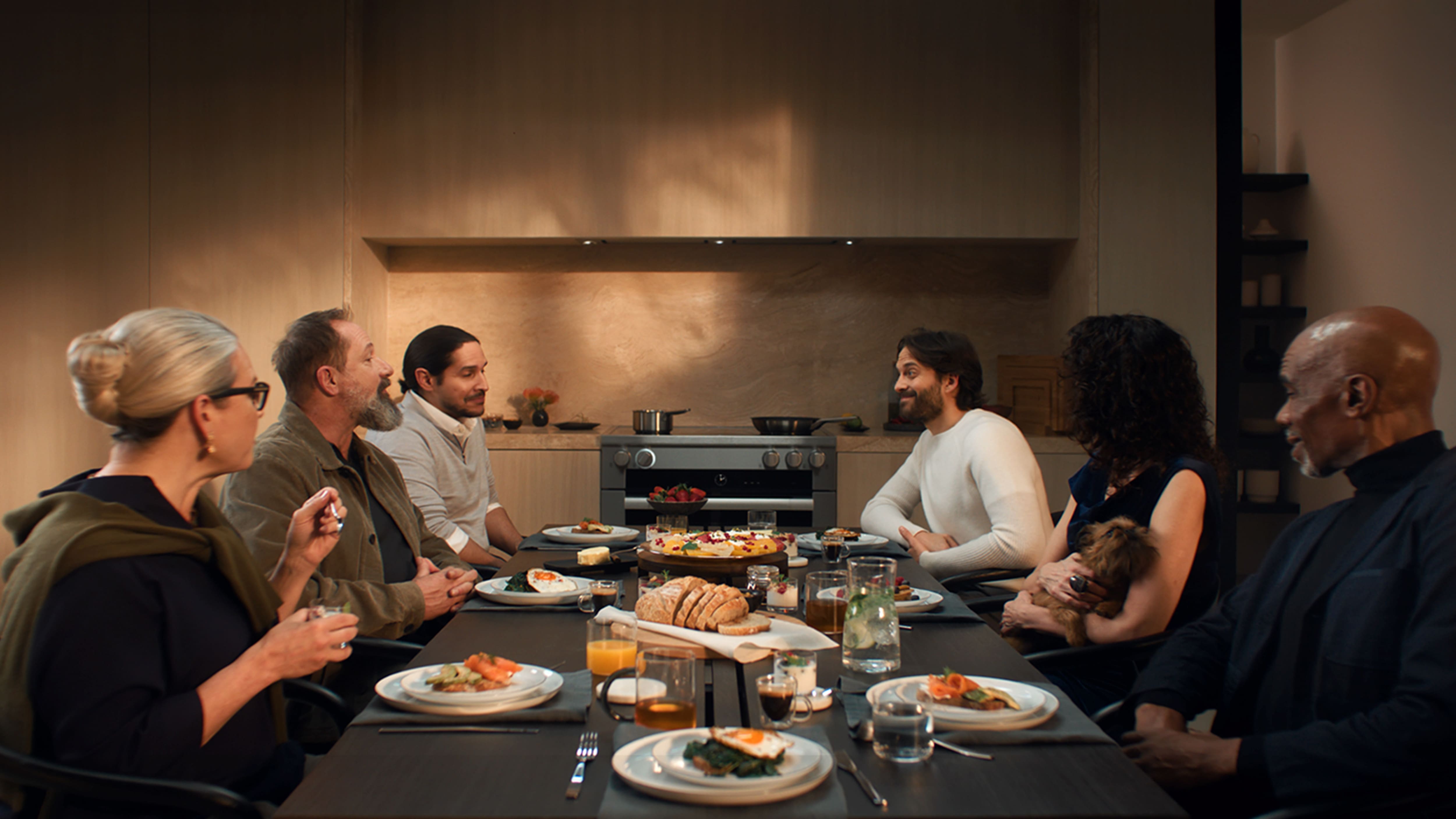 Group gathered at a modern kitchen table for brunch, with eggs, bread, fruit, and drinks; one guest gently holding a small dog. Group gathered at a modern kitchen table for brunch, with eggs, bread, fruit, and drinks; one guest gently holding a small dog.