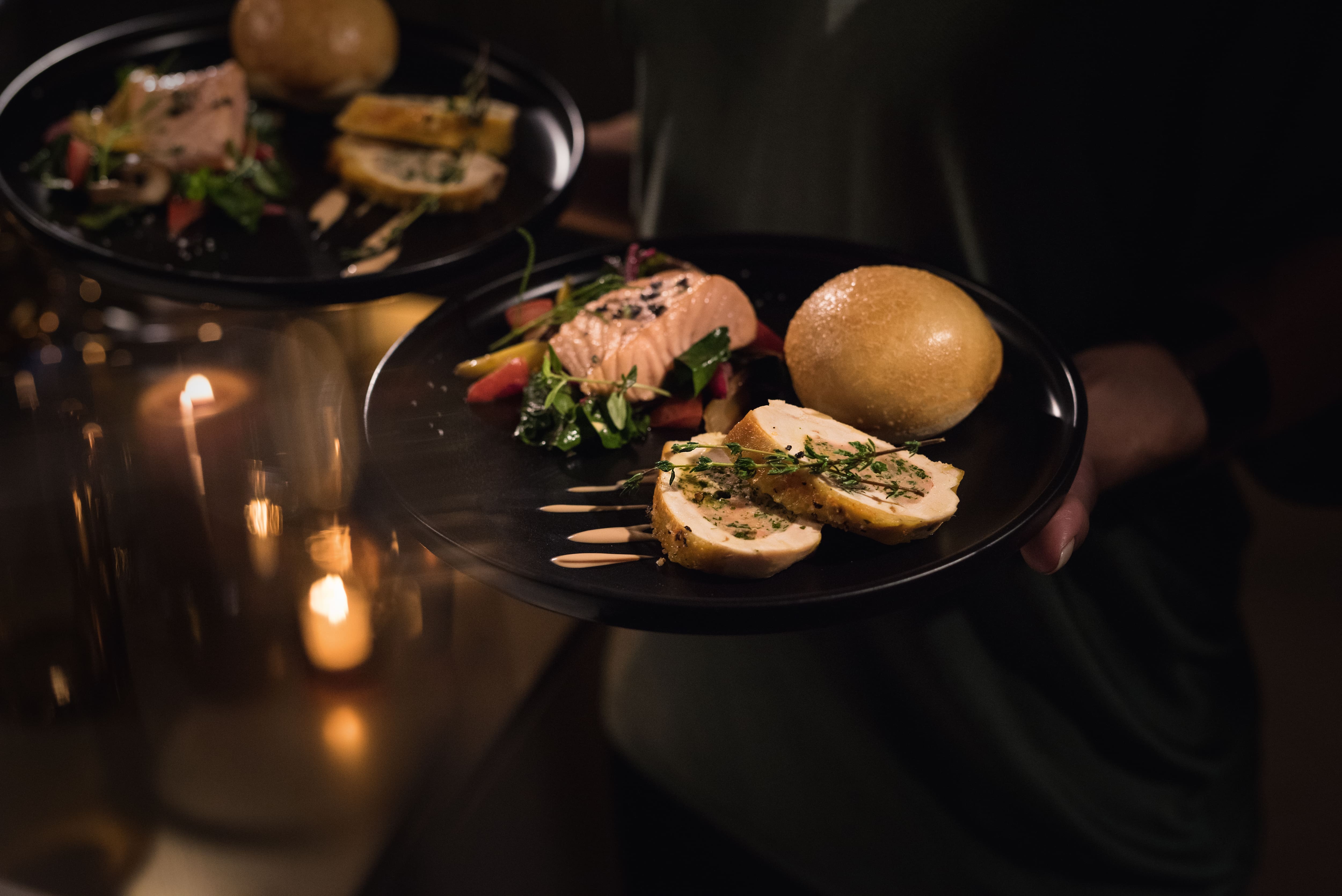 Close-up of plated gourmet meal: sliced chicken, salmon, mixed greens, and a bread roll in a candlelit setting.