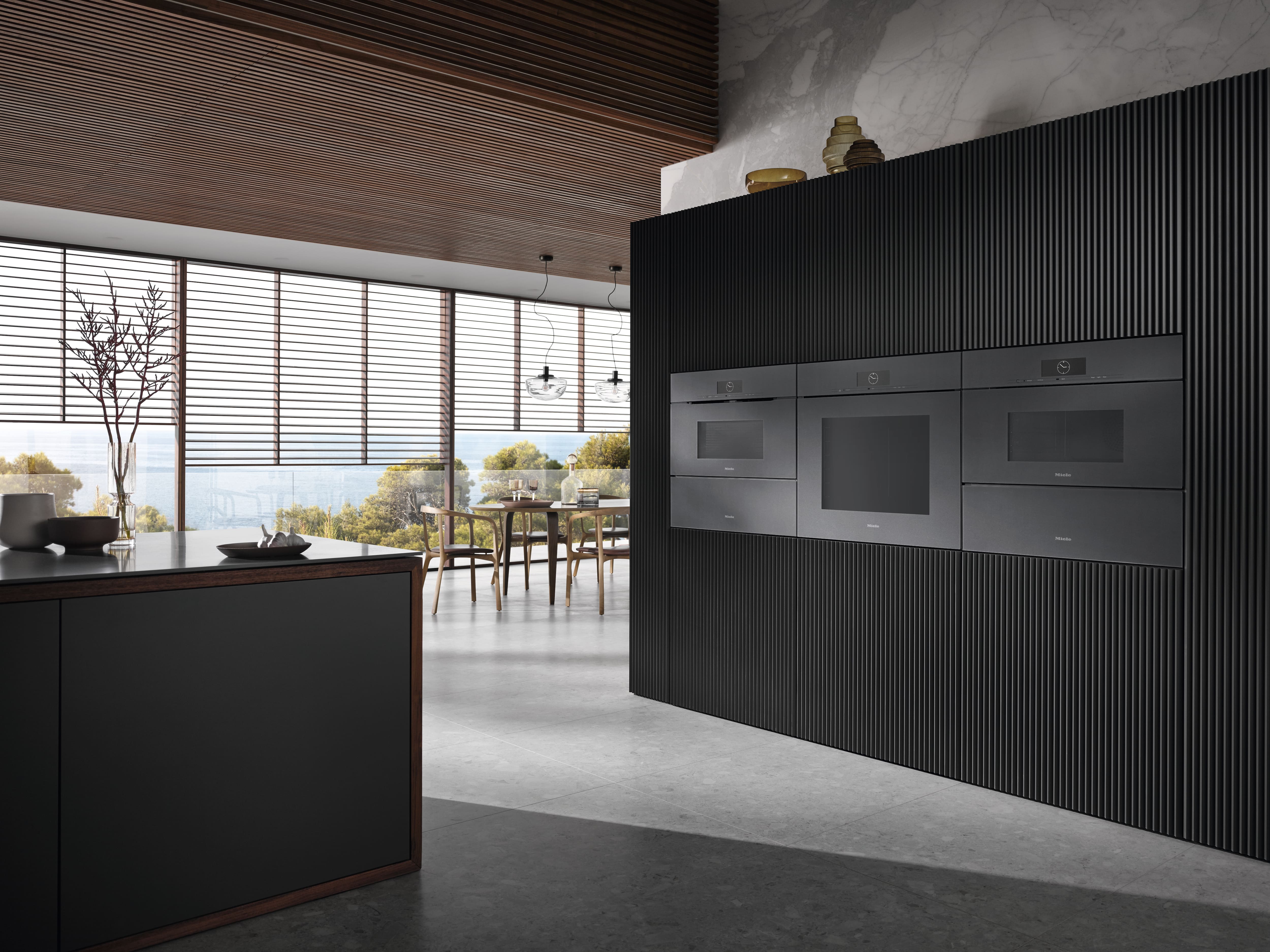 Cocina minimalista con hornos empotrados oscuros, pared estriada, isla y ventanales con vista a los árboles y al mar.