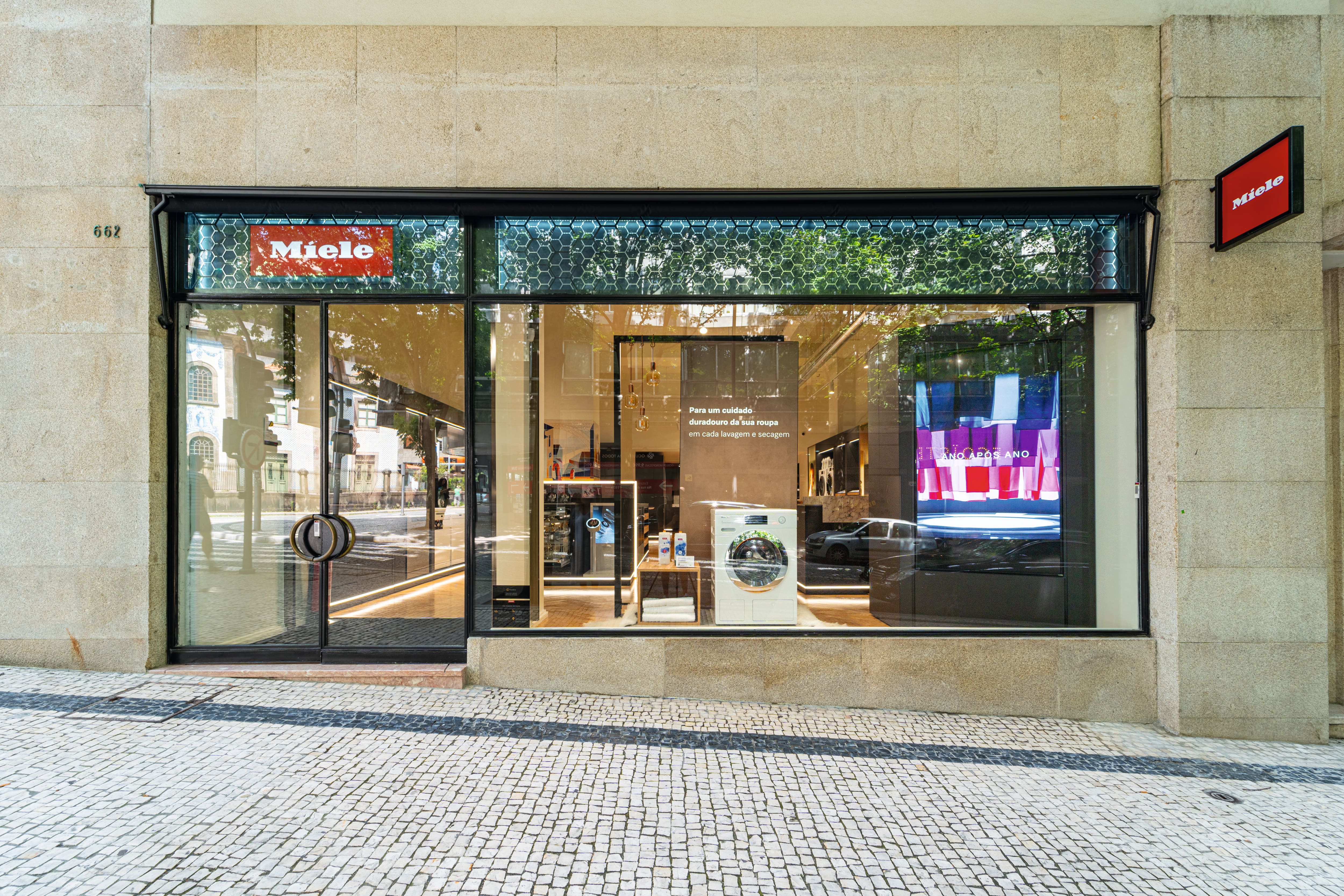 Vista de rua de uma loja do MEC Porto com grandes janelas de vidro exibindo uma máquina de lavar e interior moderno. Vista de rua de uma loja do MEC Porto com grandes janelas de vidro exibindo uma máquina de lavar e interior moderno.