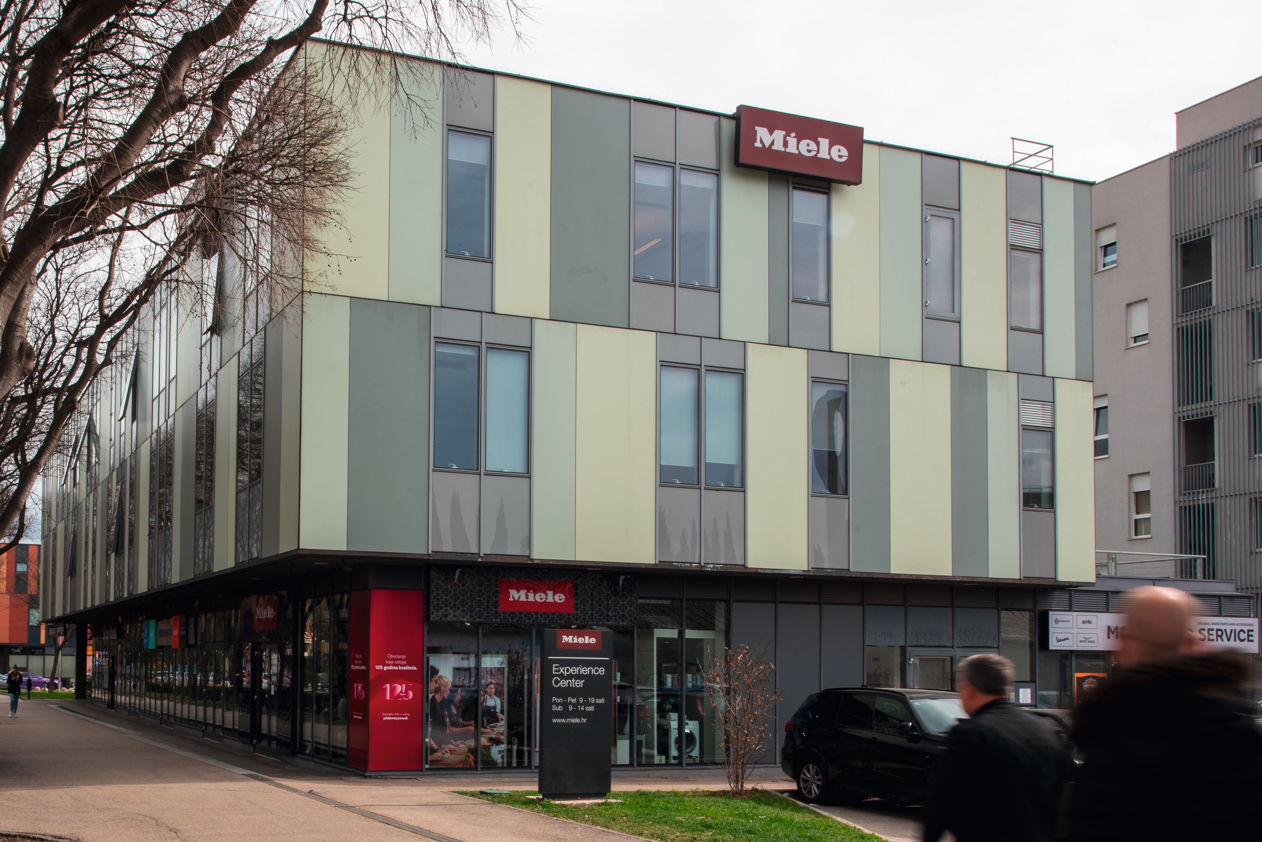 Moderna višekatnica s Miele Experience Center izlogom; prolaznici prolaze, a automobil je parkiran. Moderna višekatnica s Miele Experience Center izlogom; prolaznici prolaze, a automobil je parkiran.