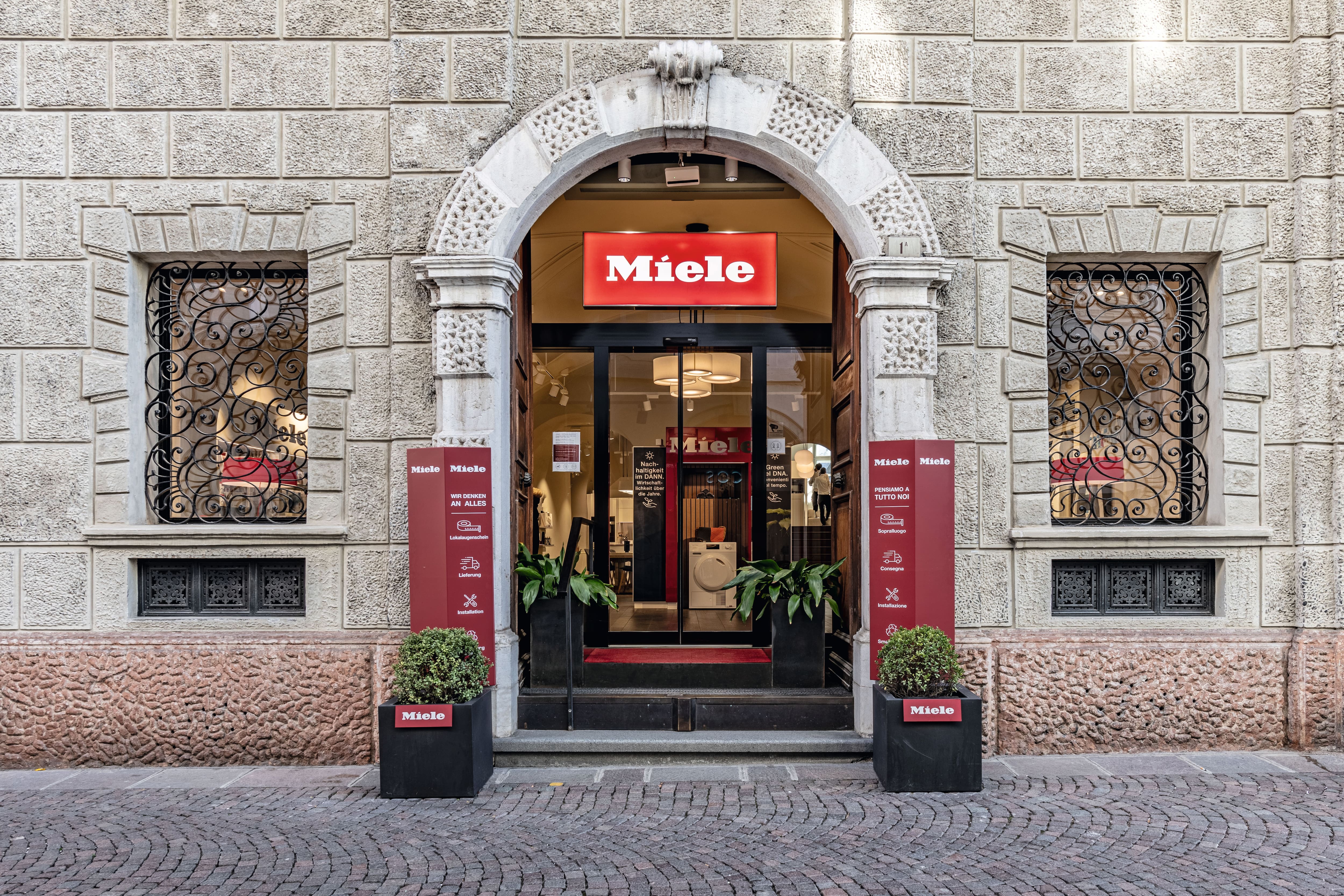 Vista della strada di un negozio Miele con facciata in pietra ad arco, insegna rossa, porte in vetro e piante in vaso. Vista della strada di un negozio Miele con facciata in pietra ad arco, insegna rossa, porte in vetro e piante in vaso.