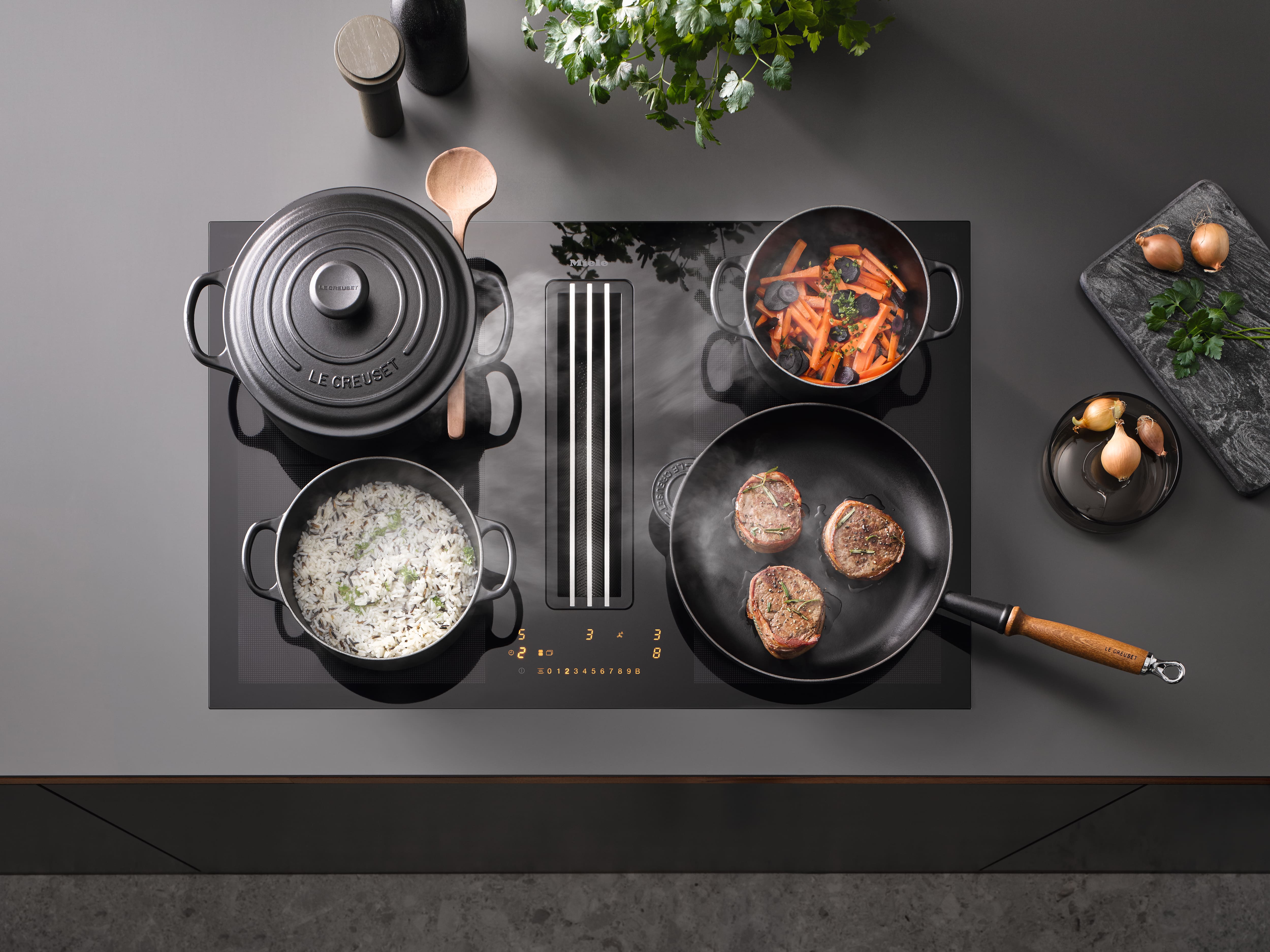 Top-down view of a Miele induction cooktop with downdraft vent, pots and pan cooking rice, carrots, and searing steaks. Top-down view of a Miele induction cooktop with downdraft vent, pots and pan cooking rice, carrots, and searing steaks.