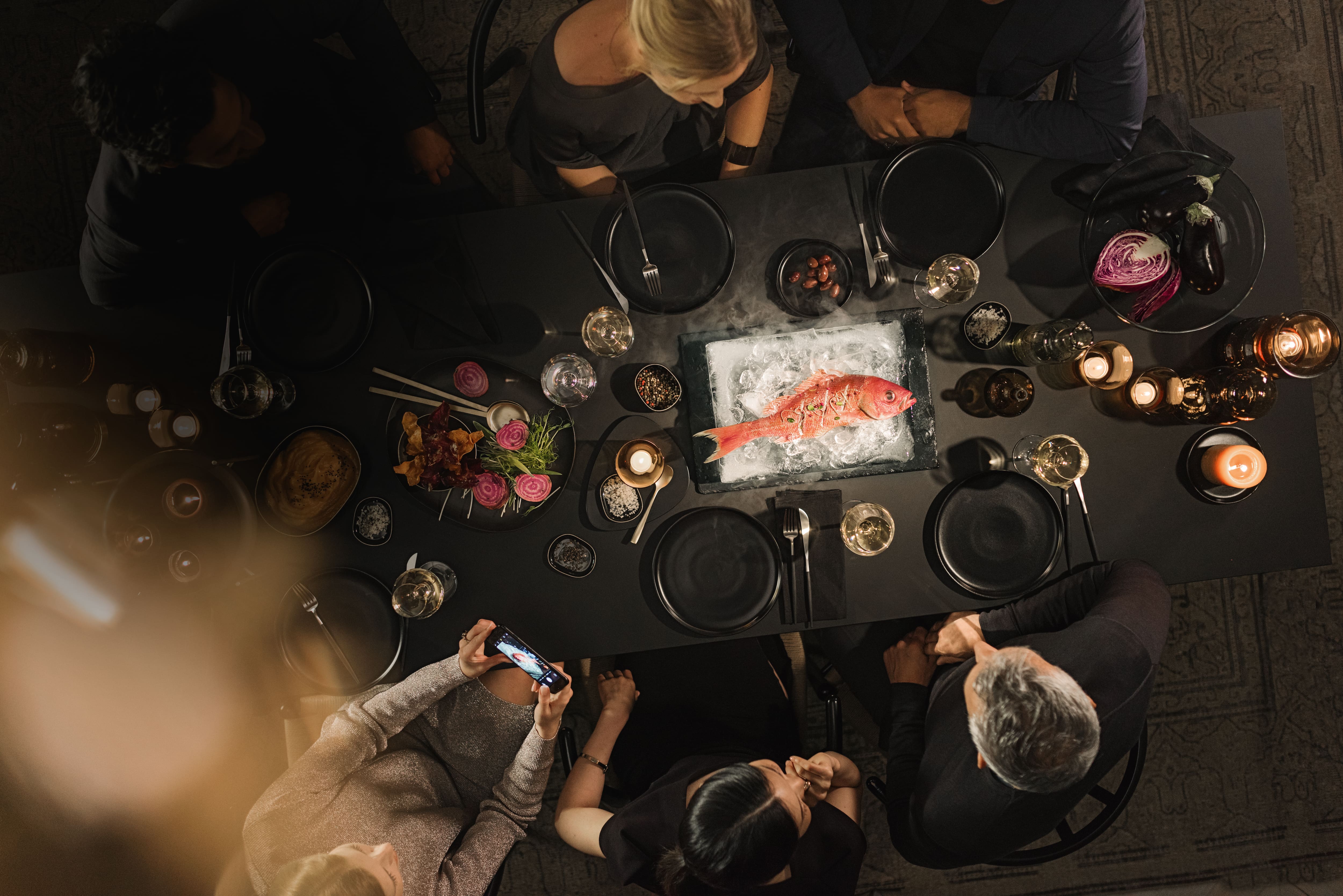 Top-down view of a moody dinner table with guests, candles, and a whole red fish on ice as centerpiece.