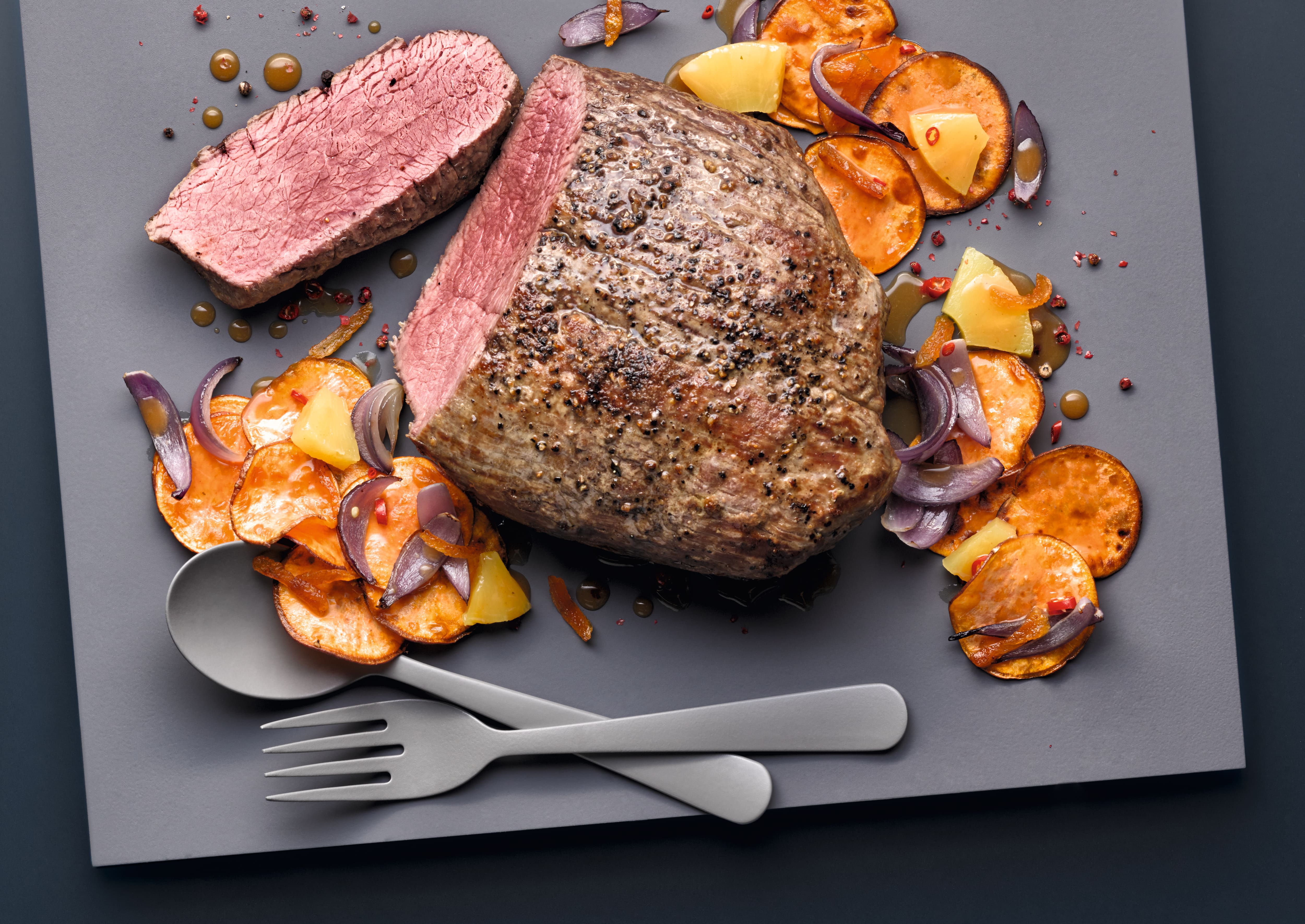 Pepper-crusted roast beef with a slice cut, served with roasted sweet potato, red onion, and sauce on a gray board. Pepper-crusted roast beef with a slice cut, served with roasted sweet potato, red onion, and sauce on a gray board.