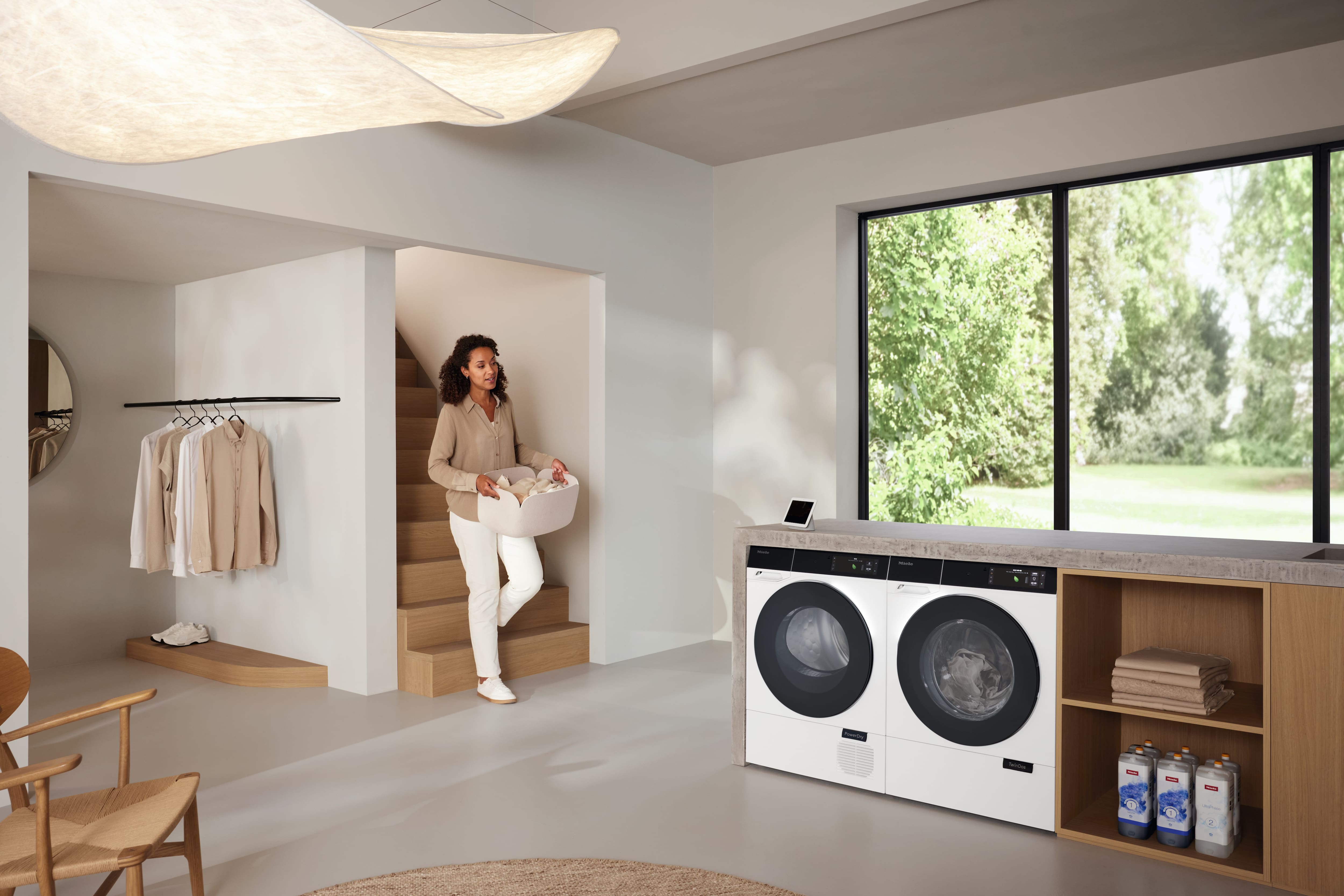 Lavadero moderno con lavadora y secadora de carga frontal; mujer llevando una cesta de ropa en un espacio luminoso y minimalista.