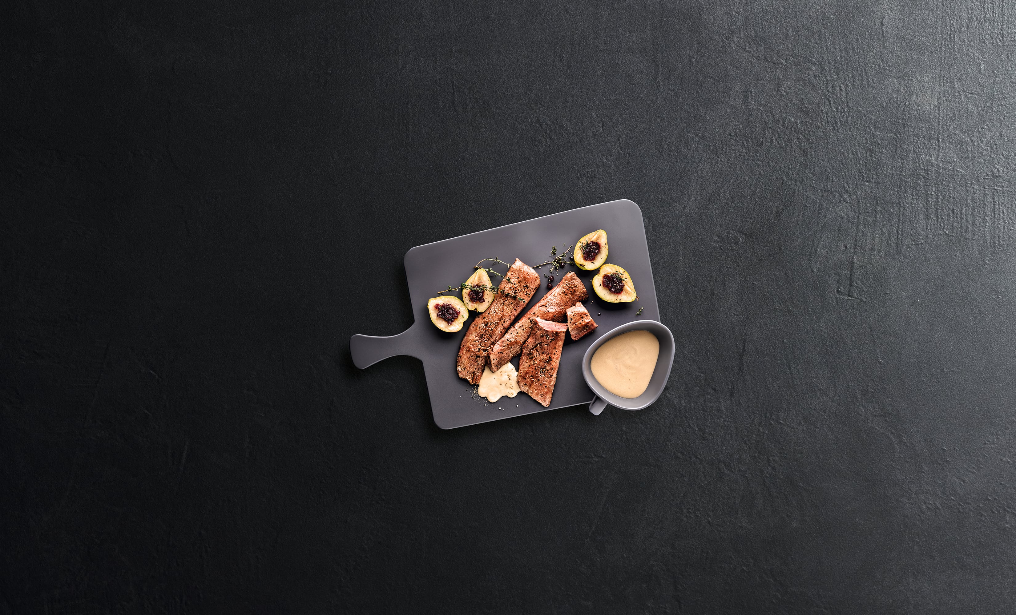 Top-down shot of sliced grilled steak with fig halves and creamy sauce on a gray board over dark surface. Top-down shot of sliced grilled steak with fig halves and creamy sauce on a gray board over dark surface.