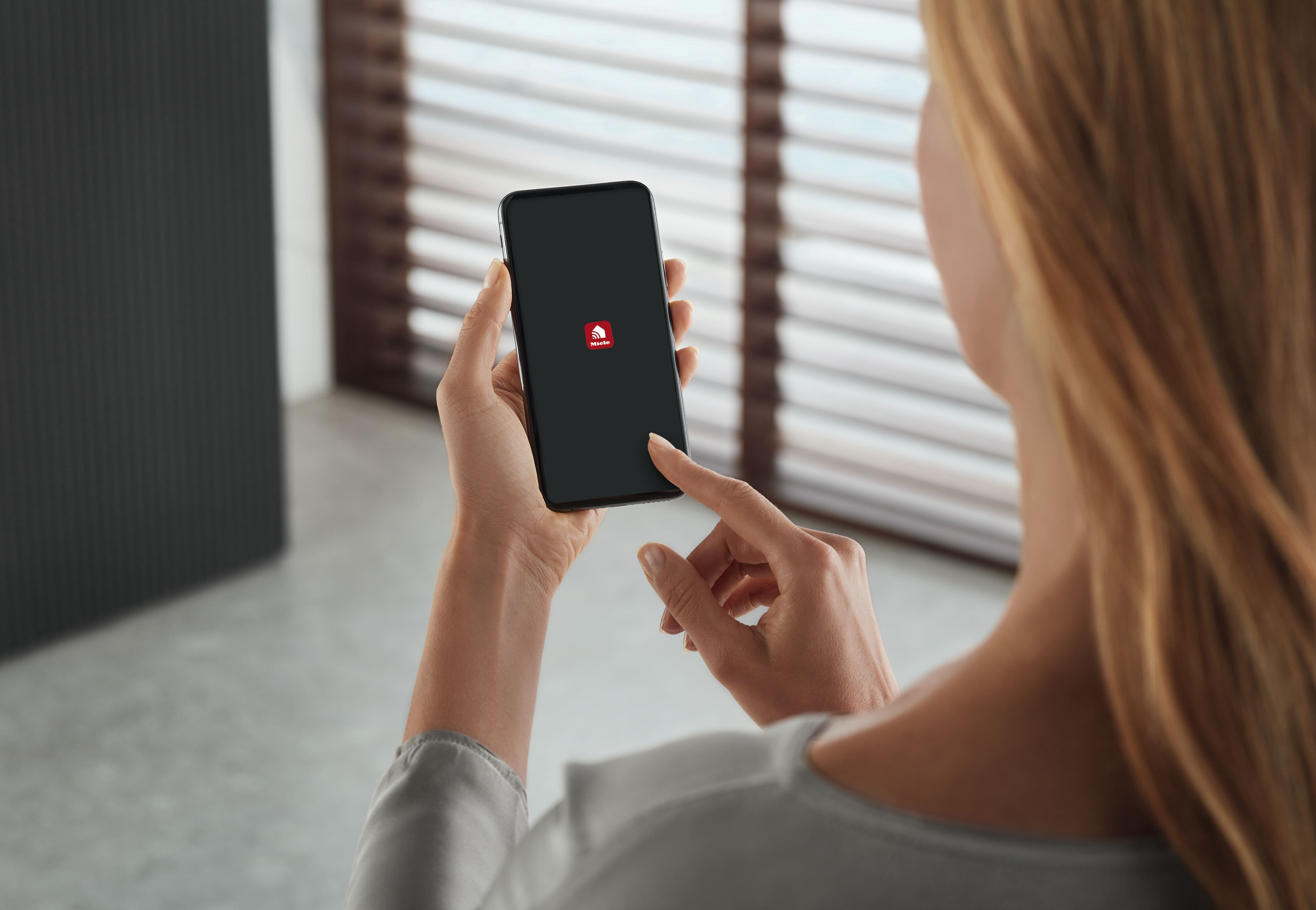 Person holding a smartphone and tapping a red app icon on a dark screen, indoors near window blinds.