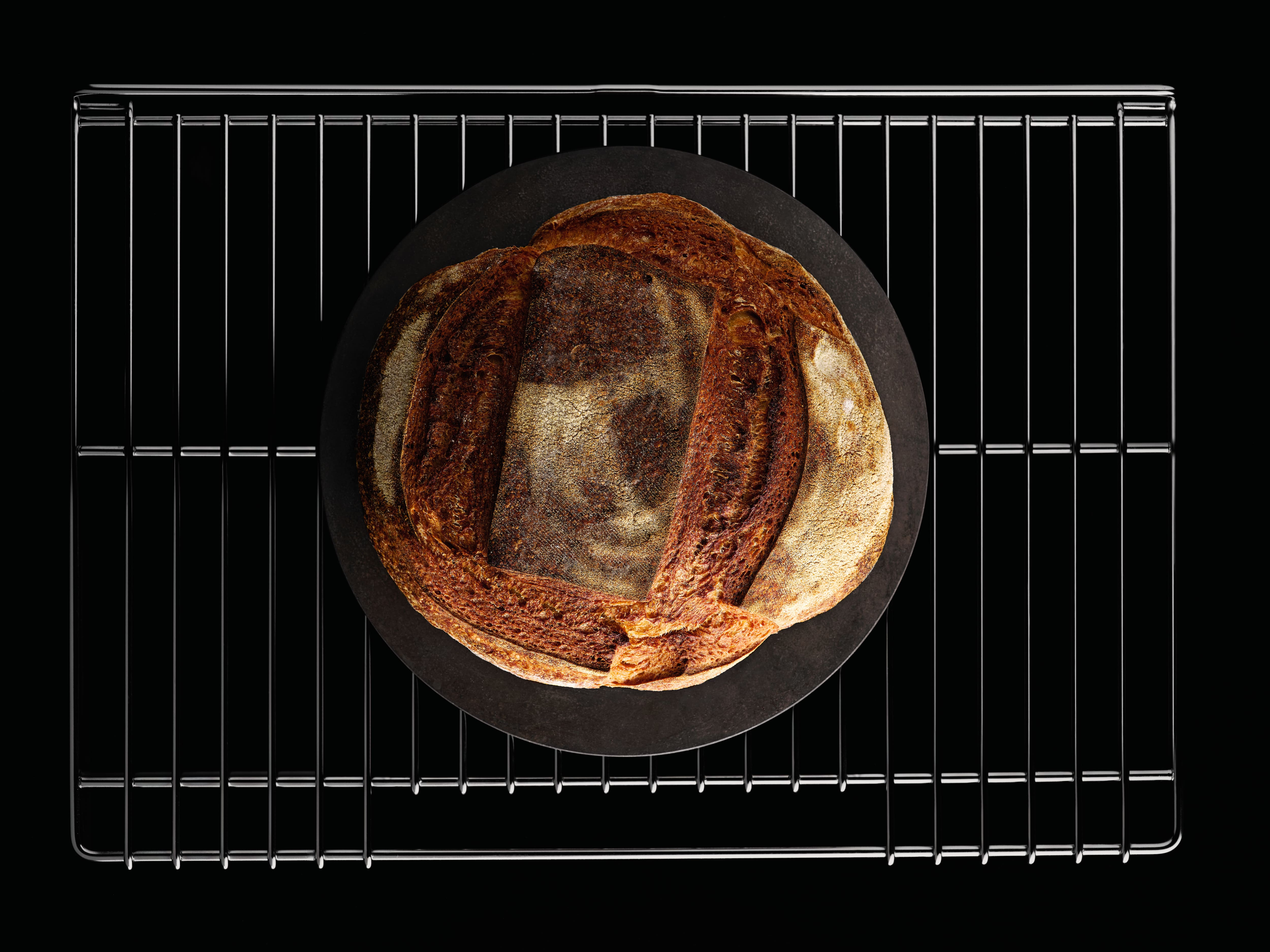 Overhead shot of a rustic sourdough loaf on a round baking stone, sitting on a metal cooling rack. Overhead shot of a rustic sourdough loaf on a round baking stone, sitting on a metal cooling rack.