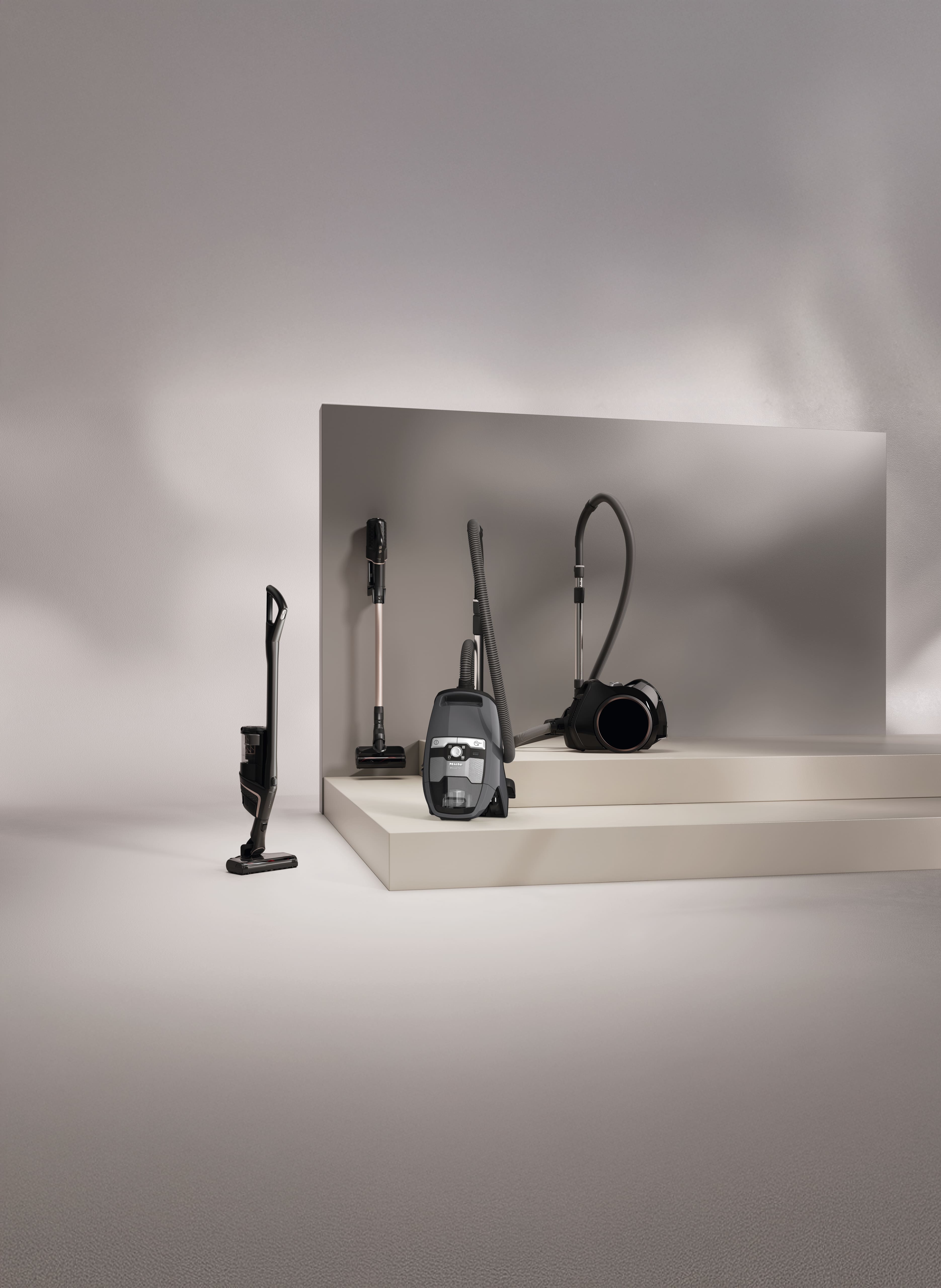 Darstellung moderner Staubsauger in einem minimalistischen Studio—kabelloser Stielstaubsauger links, ein schlanker aufrechtes Modell auf einem Ständer und ein Bodenstaubsauger mit Schlauch auf beigen Stufen vor einer grauen Wand. Darstellung moderner Staubsauger in einem minimalistischen Studio—kabelloser Stielstaubsauger links, ein schlanker aufrechtes Modell auf einem Ständer und ein Bodenstaubsauger mit Schlauch auf beigen Stufen vor einer grauen Wand.