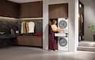 Modern laundry room with stacked washer and dryer; person loading a laundry basket on a pull-out shelf amid wood cabinetry.