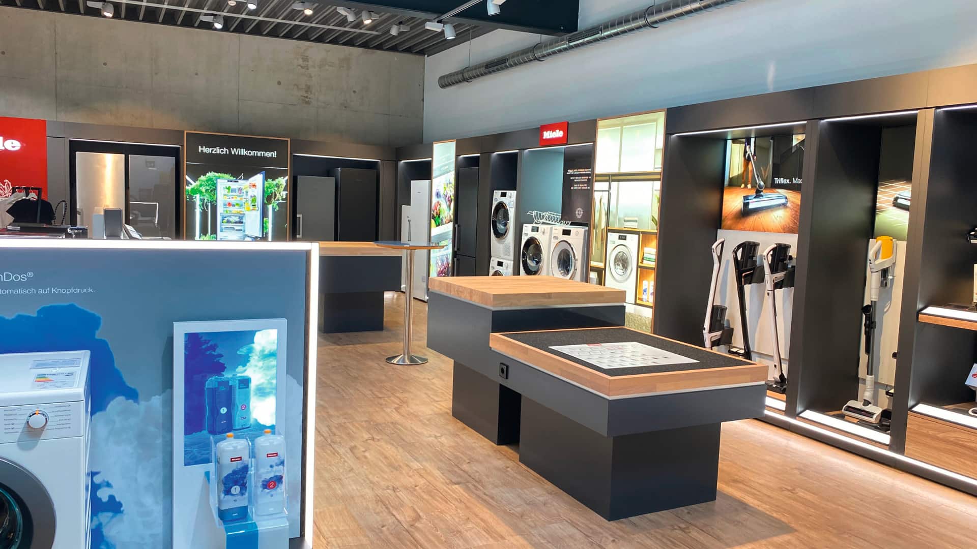 Moderner Geräteschauraum mit Miele-Branding, zeigt Kühlschränke, Waschmaschinen und kabellose Staubsauger auf Holzböden mit schwarzen Ausstellungsflächen und heller Beleuchtung.