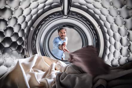 View from inside a washing machine drum as a man in a light blue shirt reaches in to load soft laundry, showcasing the interior.