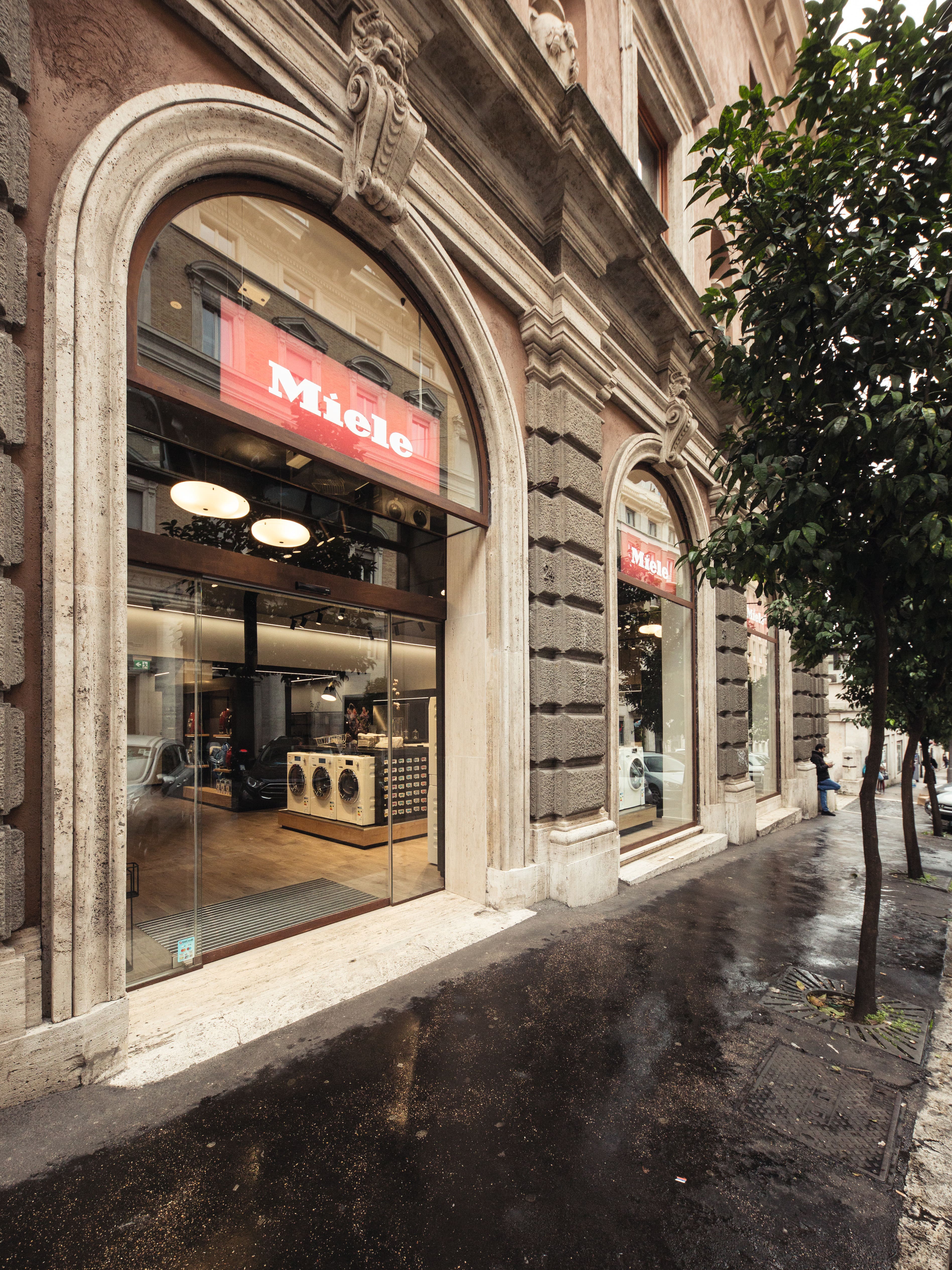 Vista della strada di uno showroom di elettrodomestici Miele in un edificio storico in pietra con alte finestre ad arco in un giorno di pioggia.