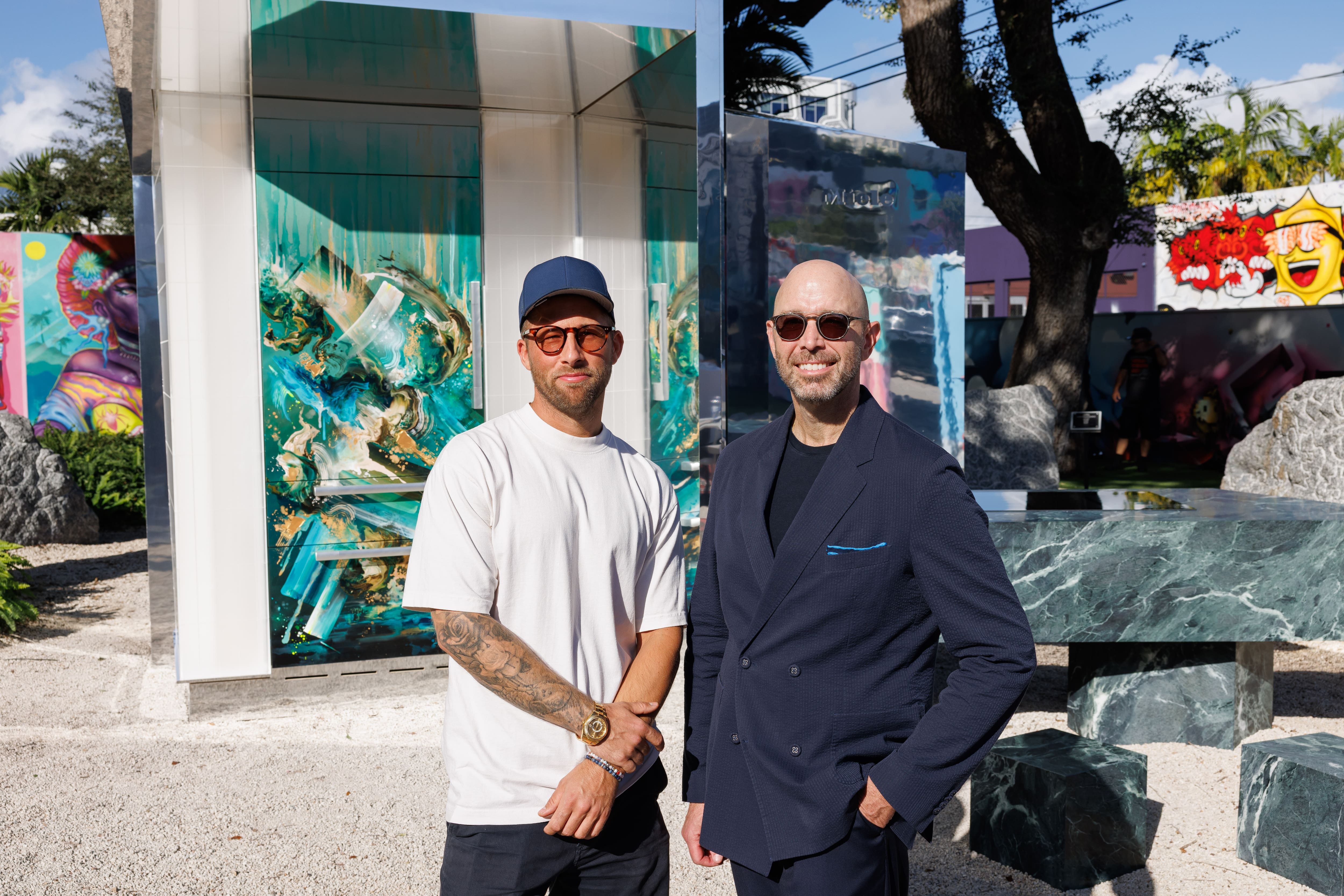 Two people pose at an outdoor art exhibit with mirrored structures, marble seating, and vibrant murals. Two people pose at an outdoor art exhibit with mirrored structures, marble seating, and vibrant murals.