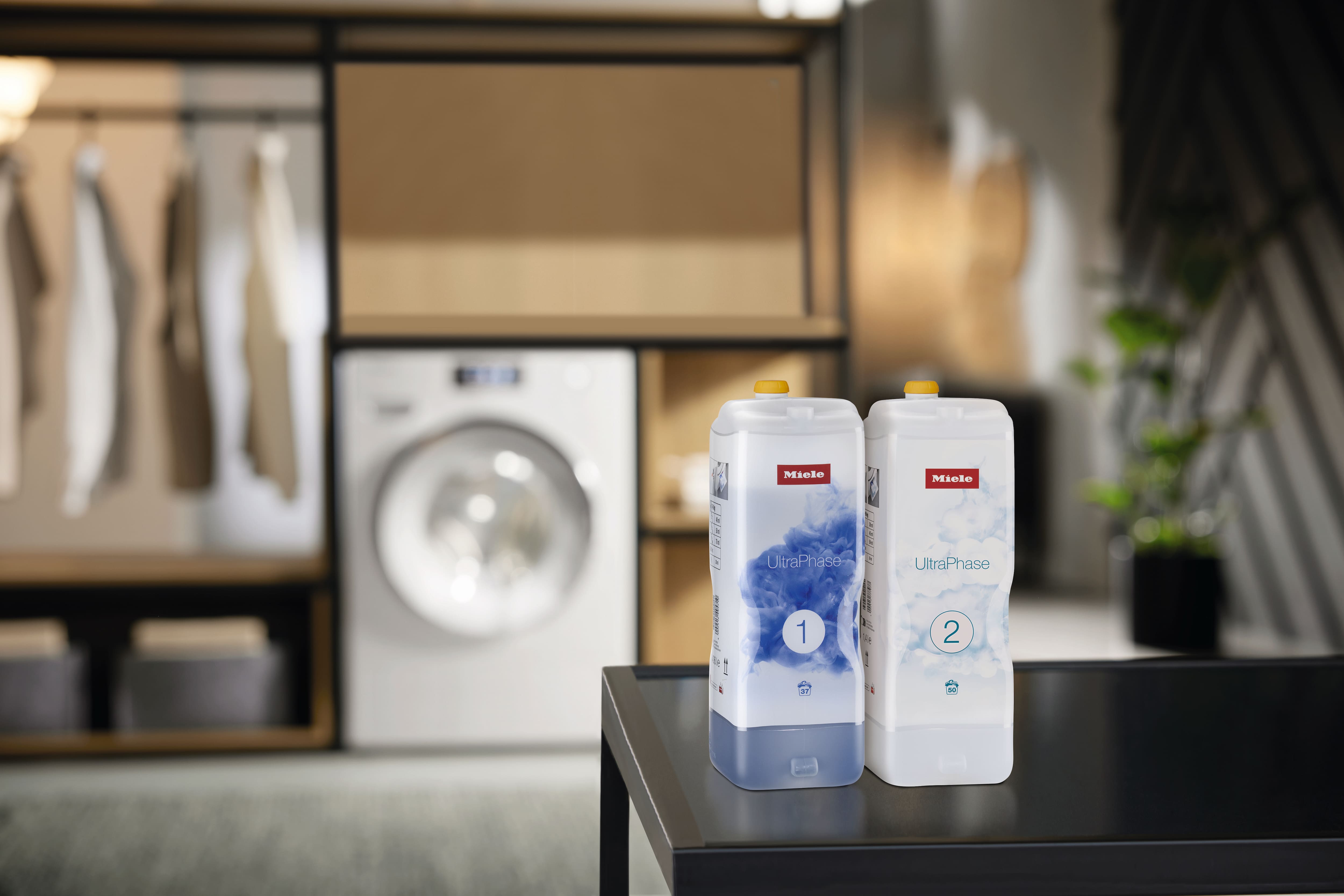 Zwei Miele UltraPhase 1 und UltraPhase 2 Waschmittelkartuschen auf einem Tisch in einem modernen Hauswirtschaftsraum; im Hintergrund ist eine Waschmaschine unscharf zu sehen.