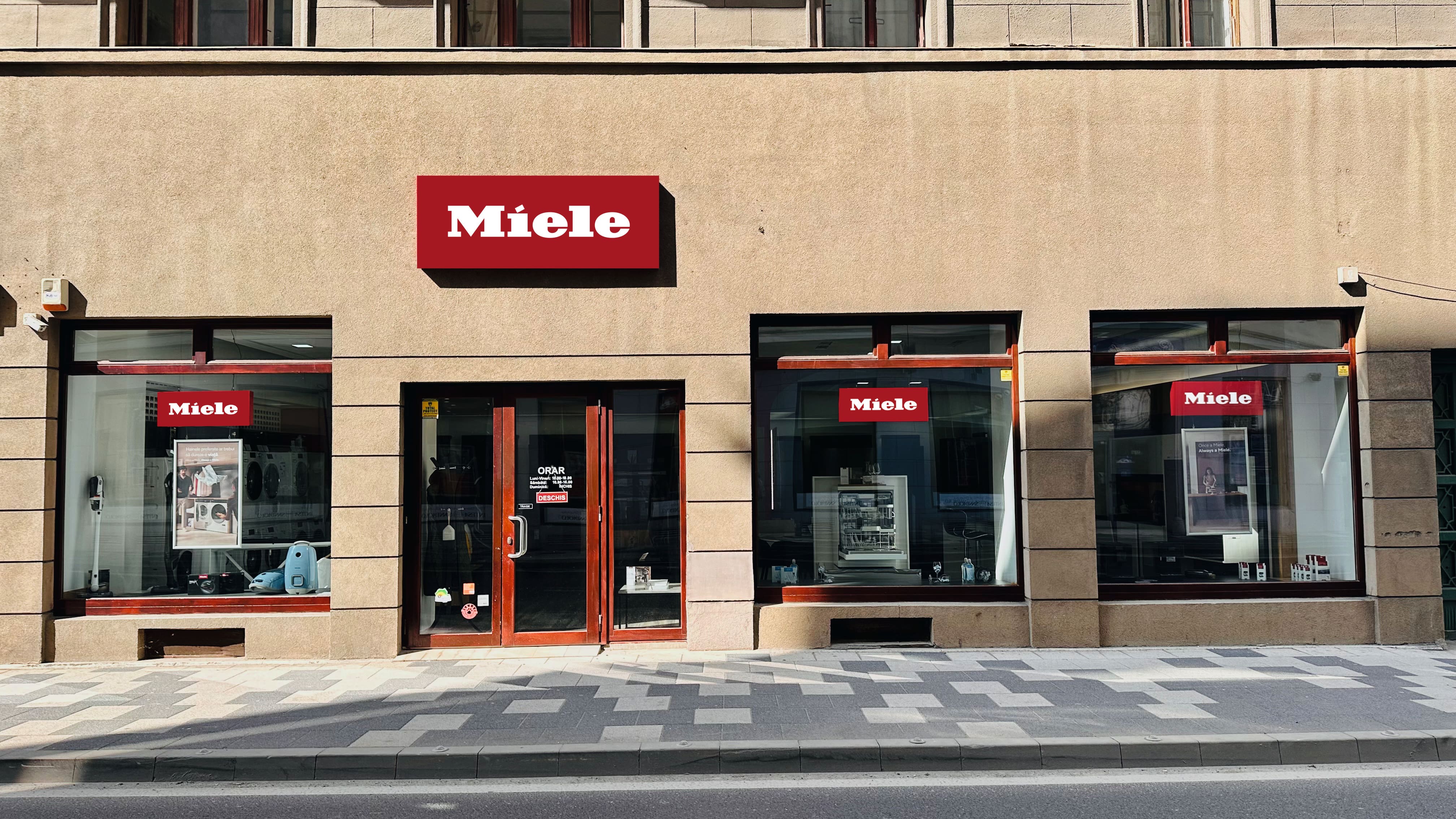 Vedere frontală a unui magazin de electrocasnice Miele cu semnalizare roșie, ferestre mari de expunere și ușă de sticlă. Vedere frontală a unui magazin de electrocasnice Miele cu semnalizare roșie, ferestre mari de expunere și ușă de sticlă.