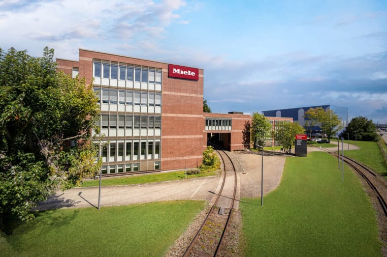 Aussenansicht eines modernen Miele Büro- und Fabrikgebäudes aus Backstein mit geschwungenen Bahngleisen, grünen Rasenflächen und blauem Himmel.