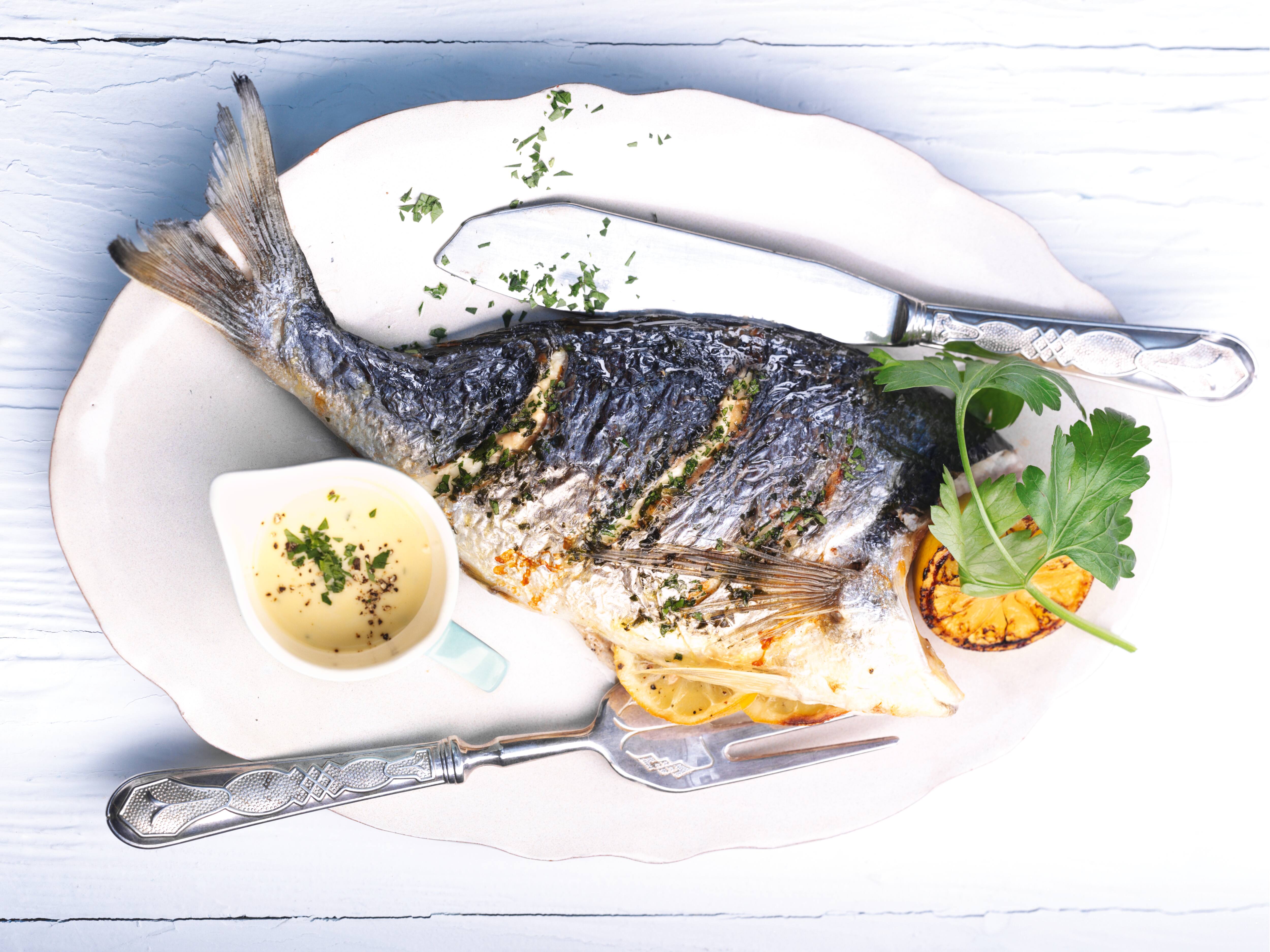 Whole grilled fish served on a platter with lemon slices, herb garnish, creamy sauce, and vintage silverware. Whole grilled fish served on a platter with lemon slices, herb garnish, creamy sauce, and vintage silverware.