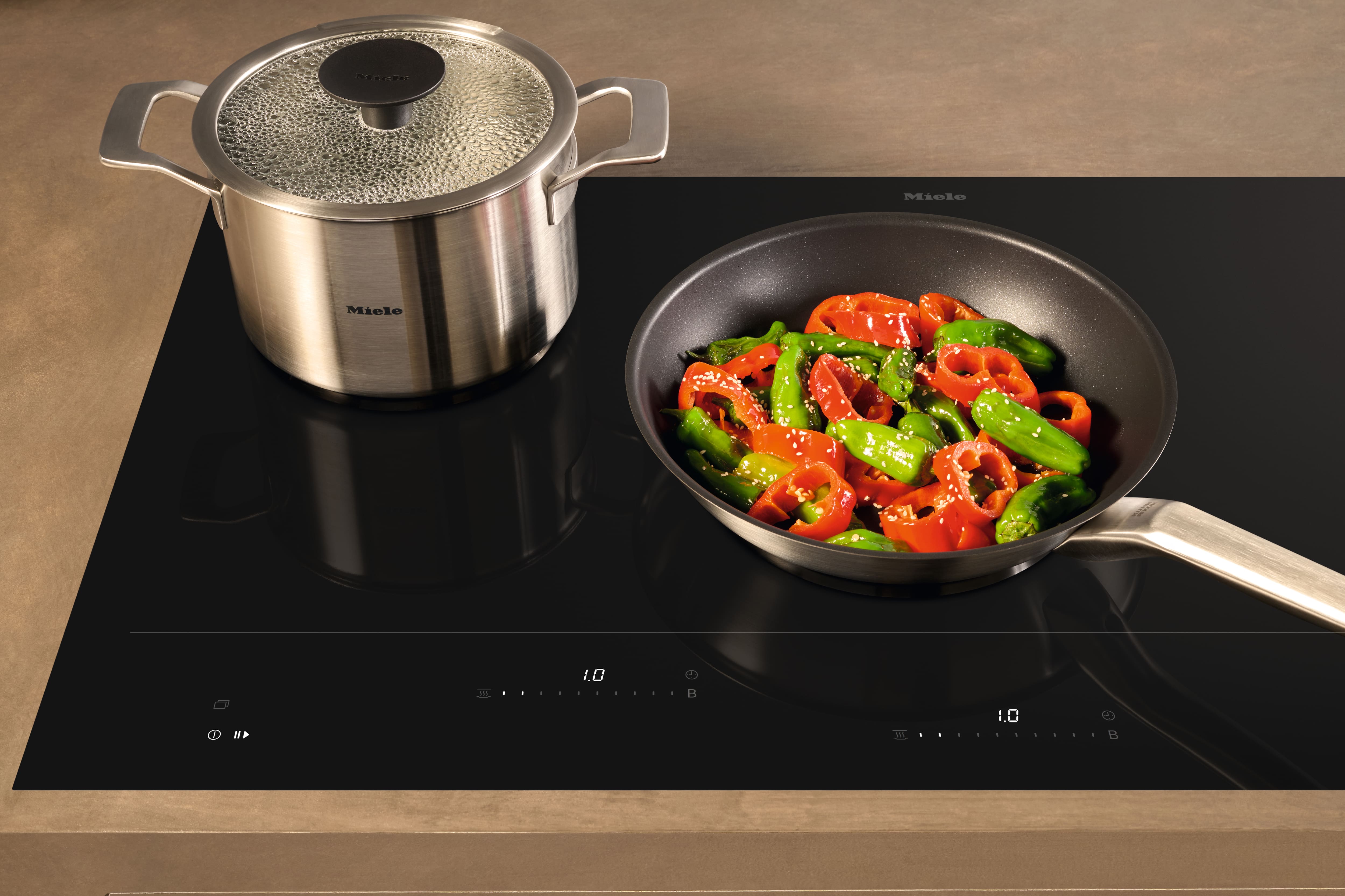 Stainless pot and frying pan with red and green peppers on a sleek Miele induction cooktop, mid-cooking. Stainless pot and frying pan with red and green peppers on a sleek Miele induction cooktop, mid-cooking.