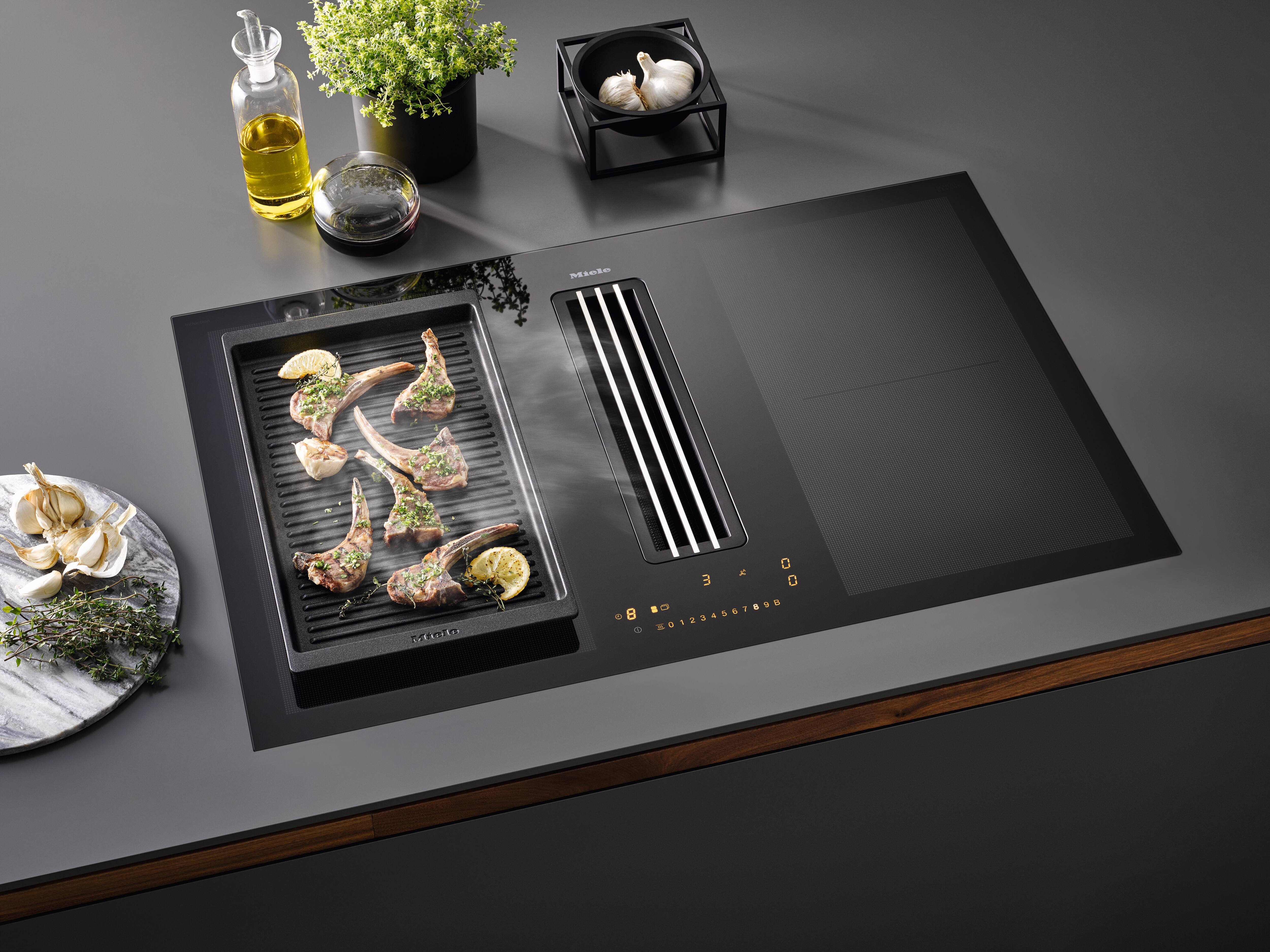 Modern induction cooktop with integrated downdraft vent and grill, searing herb-seasoned lamb chops with lemon slices. Modern induction cooktop with integrated downdraft vent and grill, searing herb-seasoned lamb chops with lemon slices.