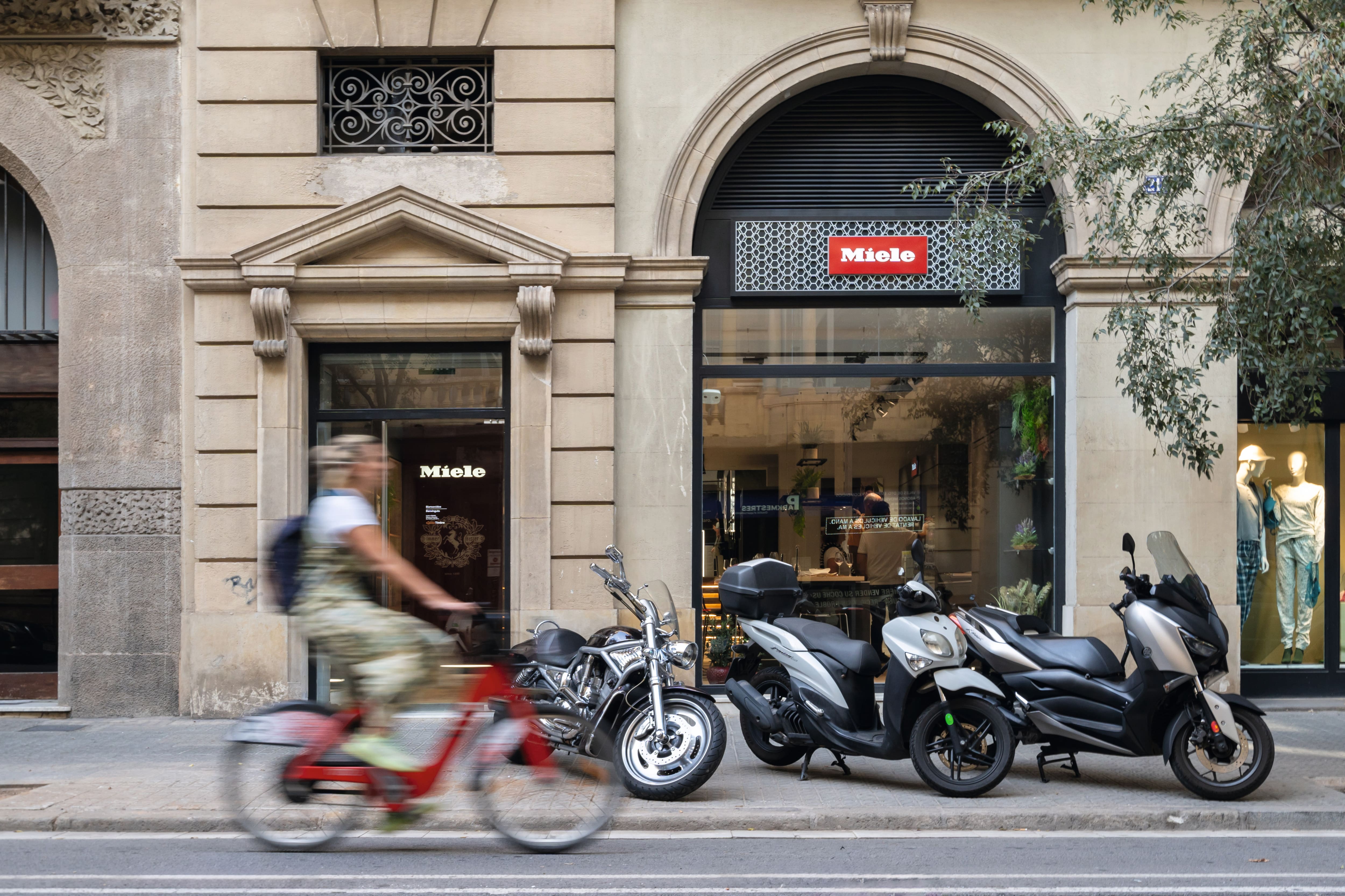 Vista de la calle de una tienda Miele con scooters estacionados y un ciclista borroso pasando, maniquíes en la ventana siguiente. Vista de la calle de una tienda Miele con scooters estacionados y un ciclista borroso pasando, maniquíes en la ventana siguiente.