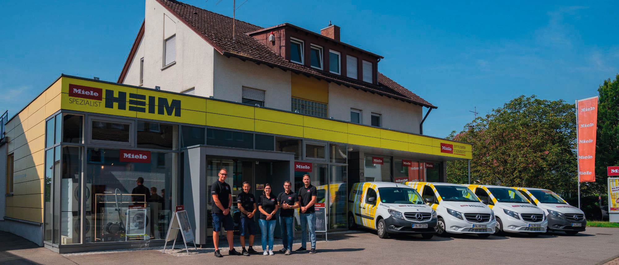 Außenansicht eines deutschen Haushaltswarengeschäfts mit gelbem „HEIM“-Schild und „Miele Spezialist“-Branding; sechs Mitarbeiter stehen davor, und vier weiße Mercedes-Servicewagen mit gelben Streifen sind an einem sonnigen Tag am Bürgersteig geparkt.