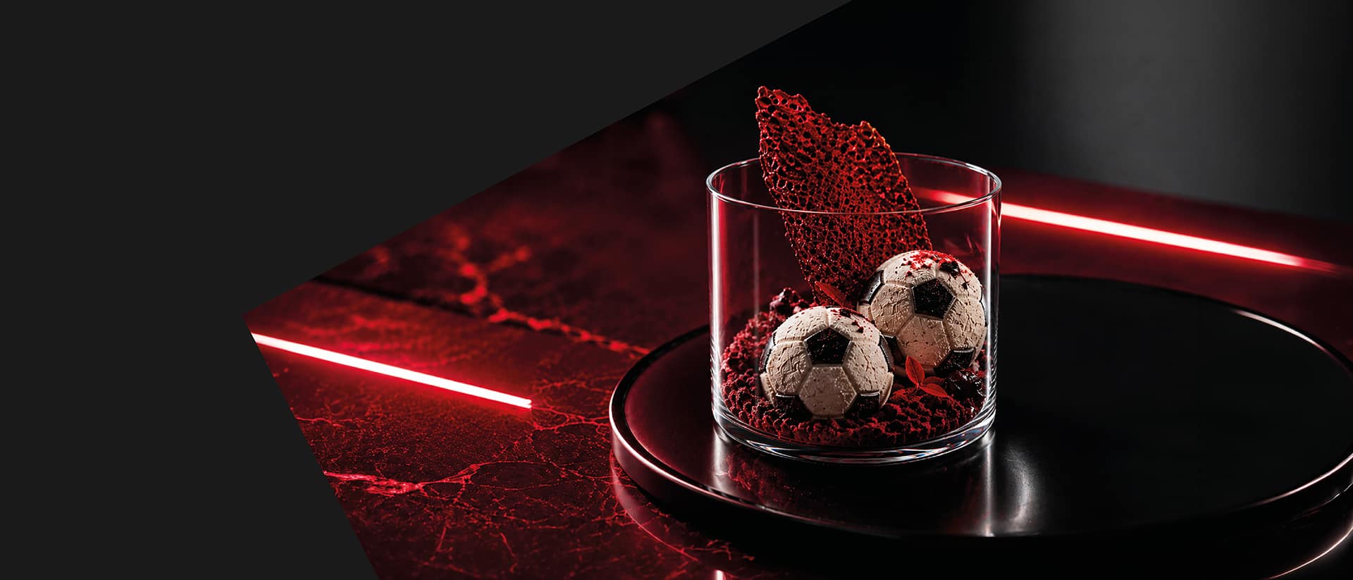 Glasdessert mit zwei schokoladenen Pralinen in Fussball-Optik auf rotem Crumble. Dramatische rote Neonbeleuchtung und schwarzer Untergrund sorgen für eine moderne, kontrastreiche Inszenierung. Glasdessert mit zwei schokoladenen Pralinen in Fussball-Optik auf rotem Crumble. Dramatische rote Neonbeleuchtung und schwarzer Untergrund sorgen für eine moderne, kontrastreiche Inszenierung.