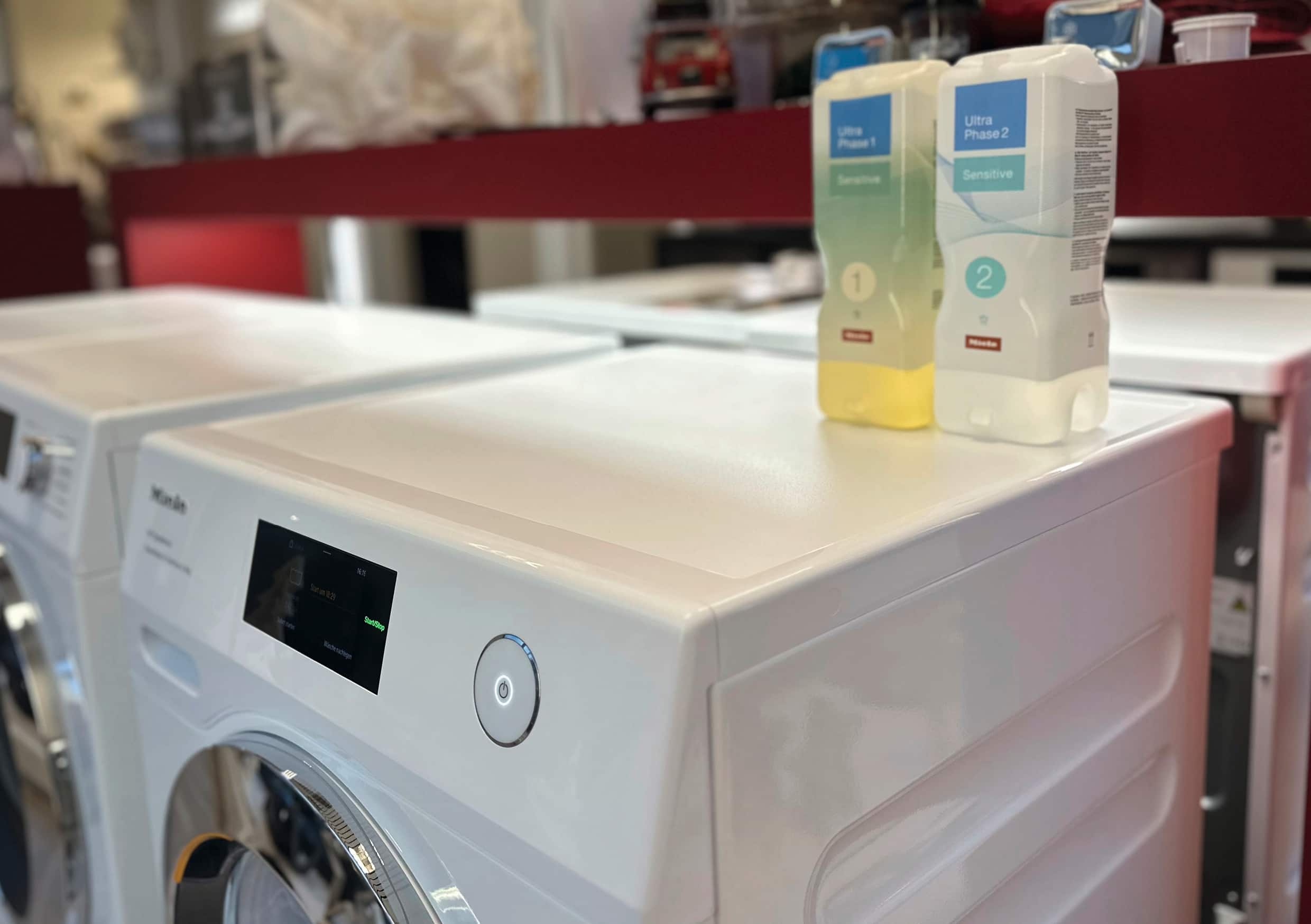 Reihe von weißen Frontlader-Waschmaschinen in einem Showroom, mit zwei Miele UltraPhase Sensitive Waschmittelflaschen (1 und 2) auf einer Maschine platziert.