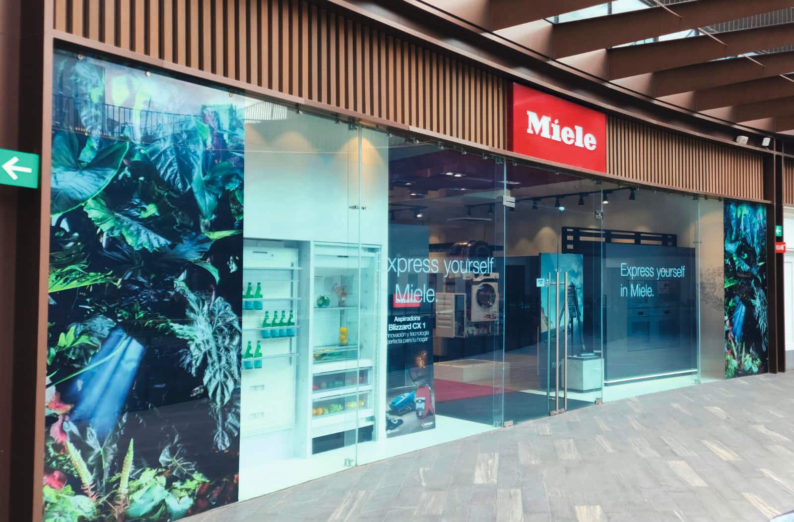 Sala de exposición de electrodomésticos Miele con frente de vidrio y letrero de 'Exprésate' con gráficos de pared con hojas en un centro comercial.